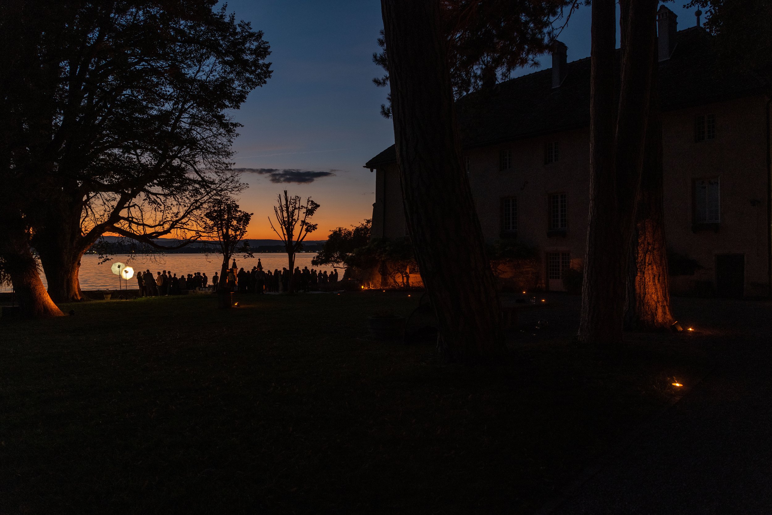 Coucher de soleil au bord de l'eau avec des silhouettes de personnes et des arbres dans une lumière tamisée, près d'un bâtiment.