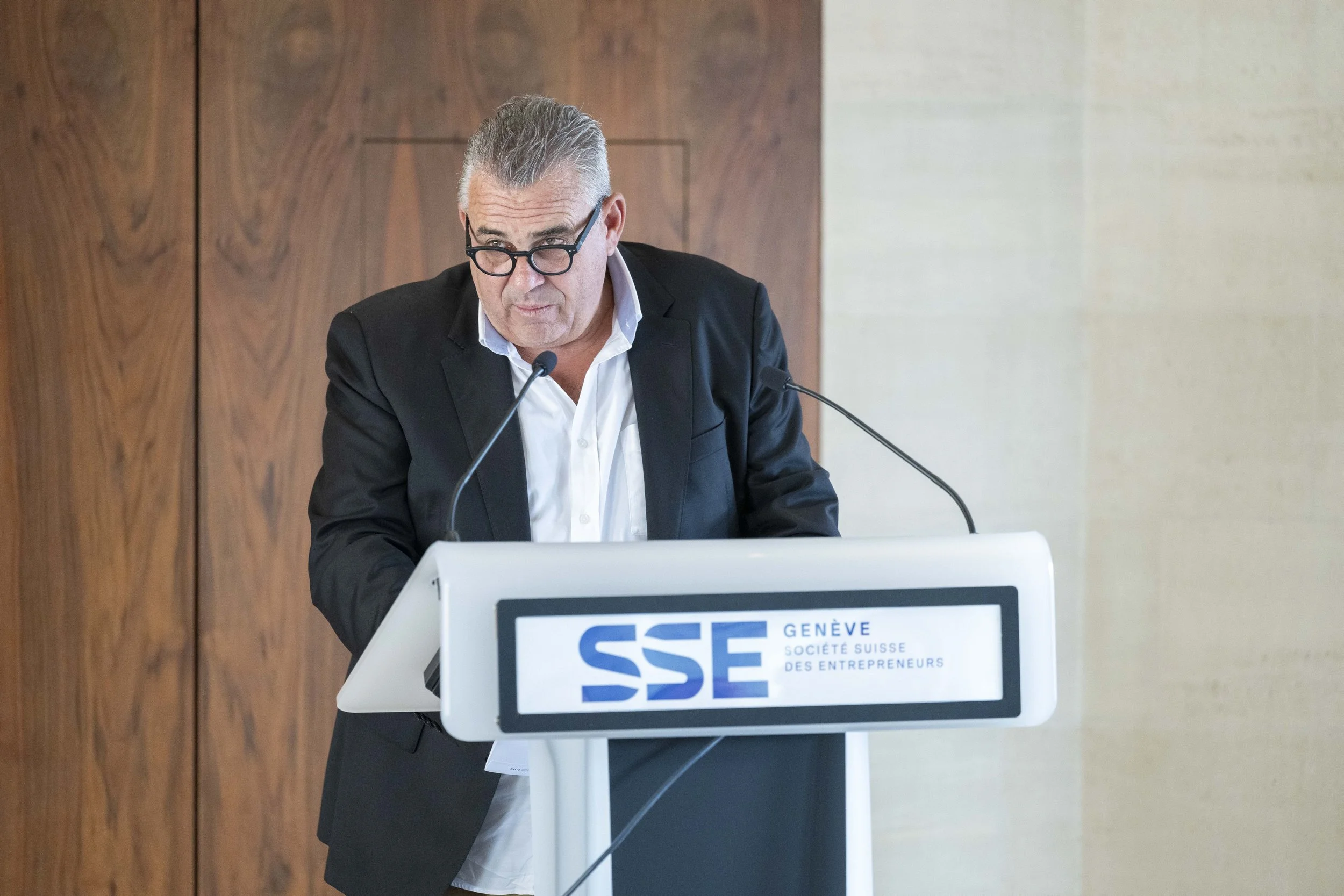 Homme portant un costume noir et lunettes, donnant une discours à la conférence, debout derrière un pupitre avec un logo SSE.