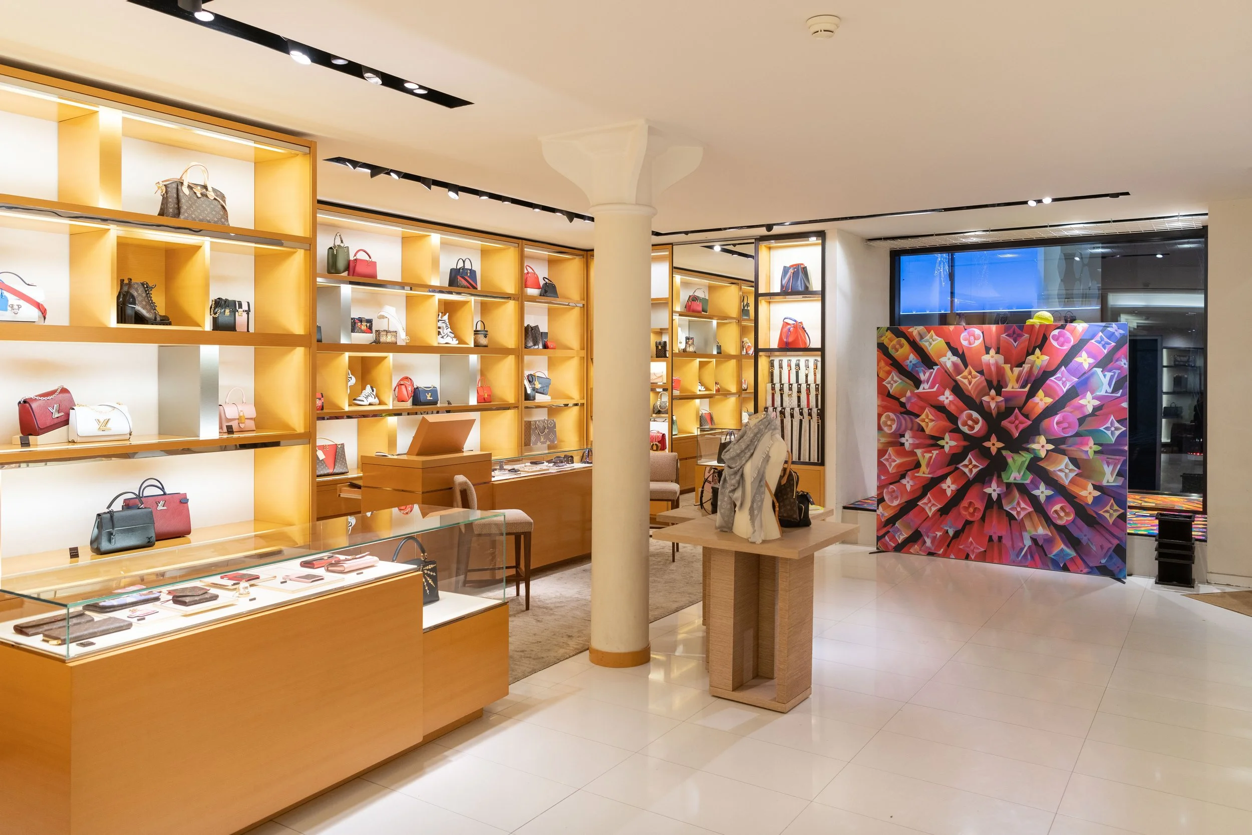 Intérieur d'une boutique de sacs à main et accessoires de luxe avec des étagères en bois remplies de sacs colorés, comptoir avec bijoux, et une installation artistique colorée en arrière-plan.