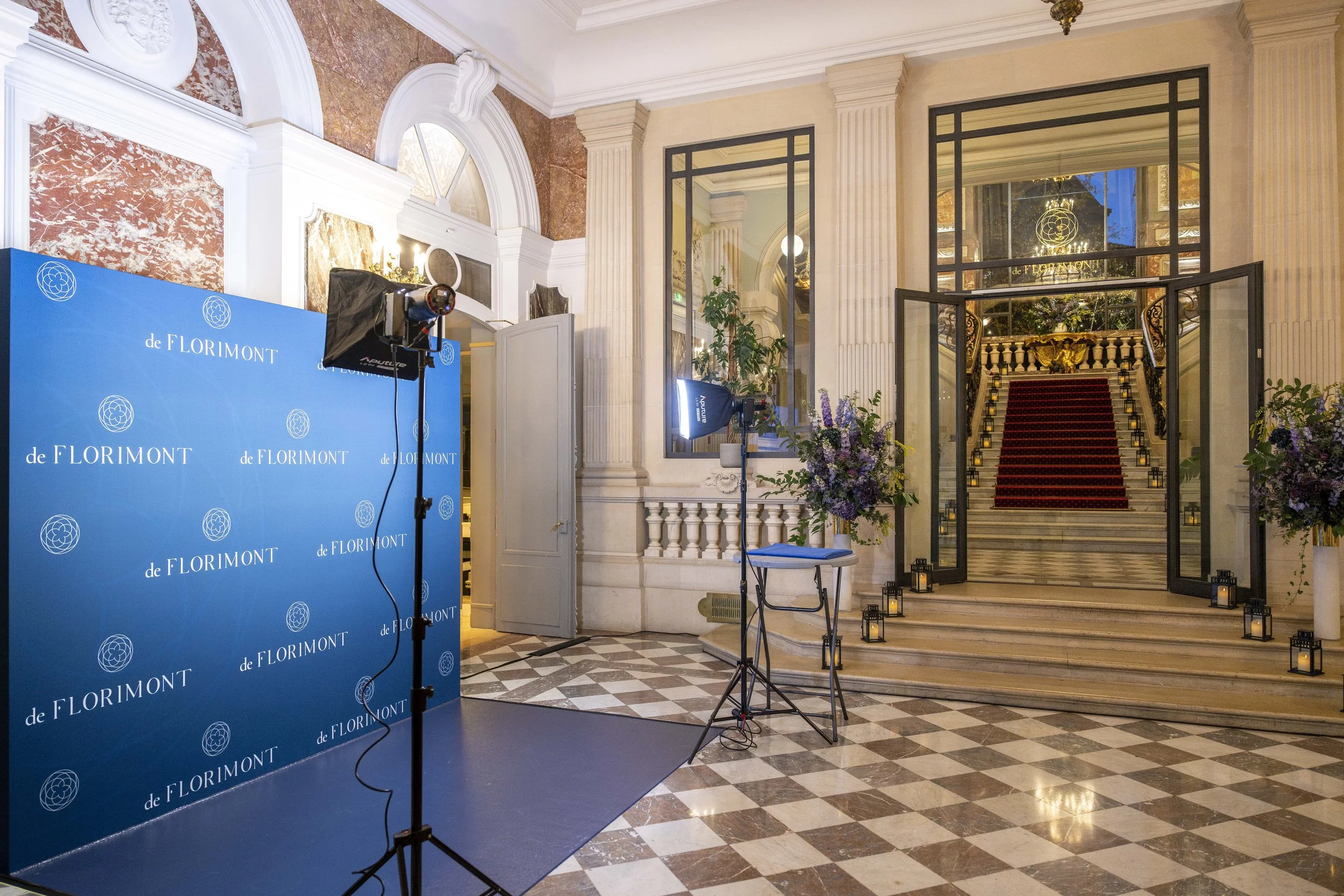 Intérieur d'un palace orné, avec un tapis rouge sur un escalier en marbre, des fleurs, des lanternes, une installation photo avec un fond bleu portant le texte 'de FLORIMONT', et un éclairage professionnel.