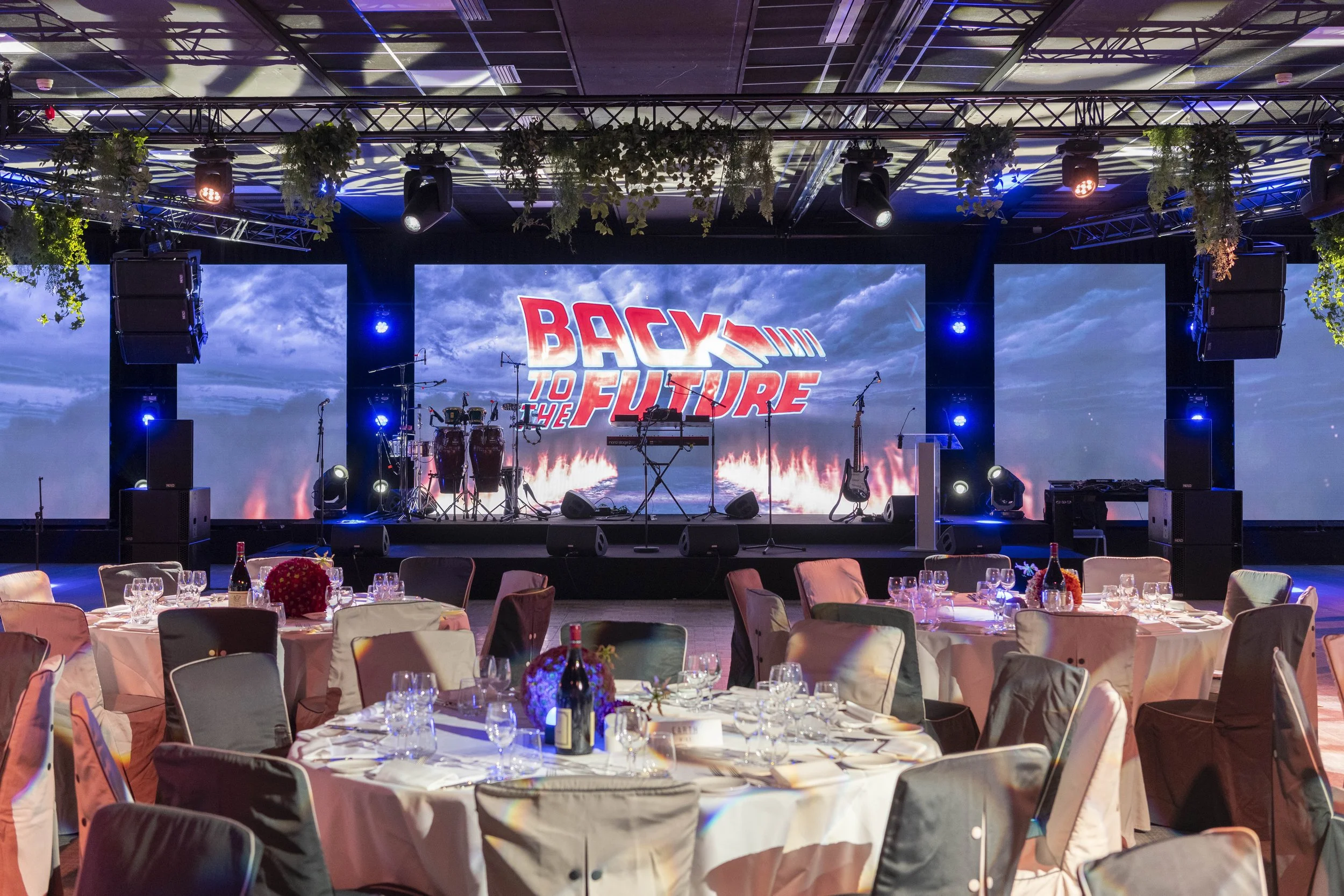 Salle de réception avec scène équipée pour un spectacle ou une conférence, écrans LED affichant 'Back to the Future', tables rondes avec nappes blanches, bouteilles de vin et décorations florales