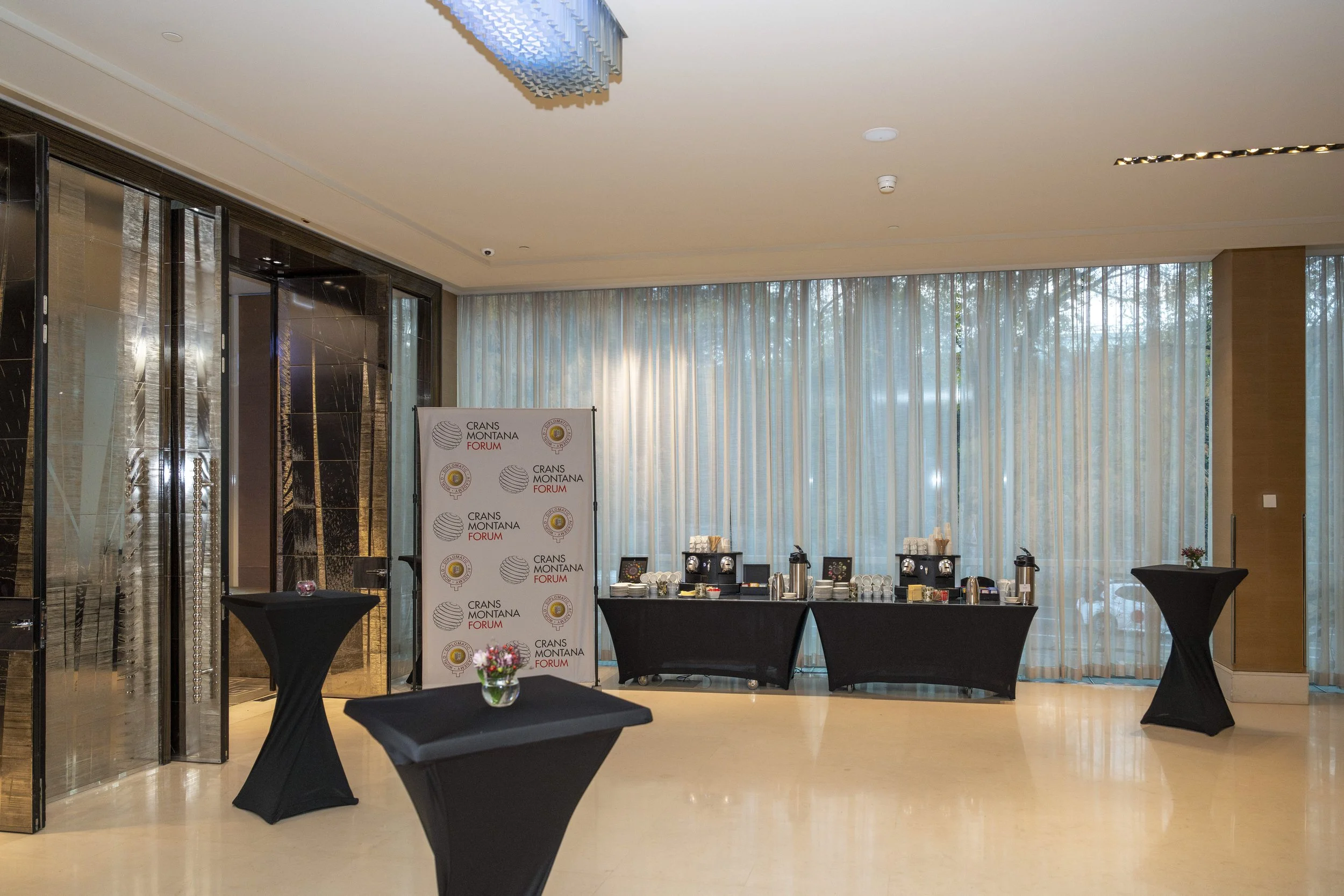 Salle de réception avec buffet, tables hautes couvertes de noir, fleurs, tapisseries en vitres, et un panneau avec logos d'un forum dans une grande pièce lumineuse.