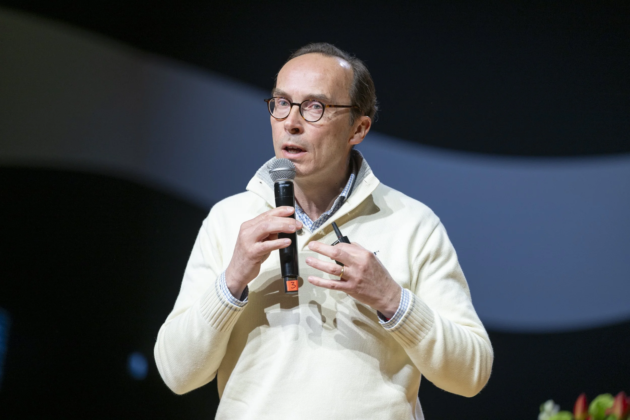 Homme parlant lors d'une conférence, tenant un microphone, portant des lunettes et un pull blanc.
