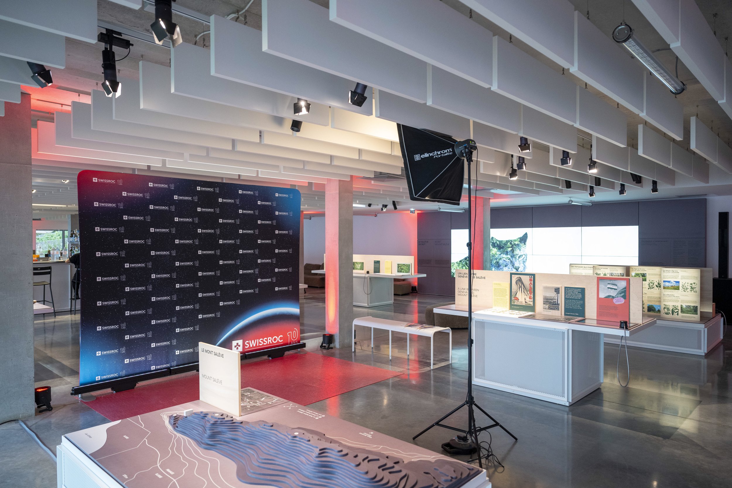 Espace d'exposition intérieur avec un fond noir affichant «SWISSROC» et «10» en bas à gauche, un écran de studio photo, un panneau d'affichage avec des textes et images, et un plafond décoré de structures blanches avec un éclairage professionnel.