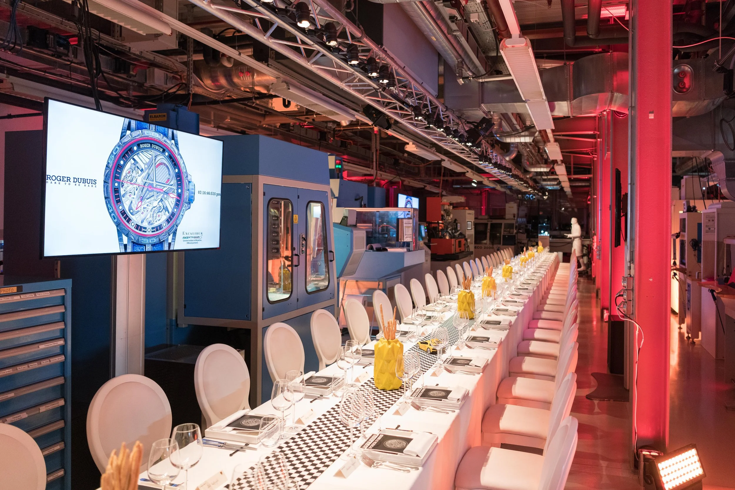 Salle de réunion avec une longue table décorée, des verres et des assiettes, entourée de chaises blanches, dans un environnement industriel éclairé en rouge, avec des écrans et des machines en arrière-plan.