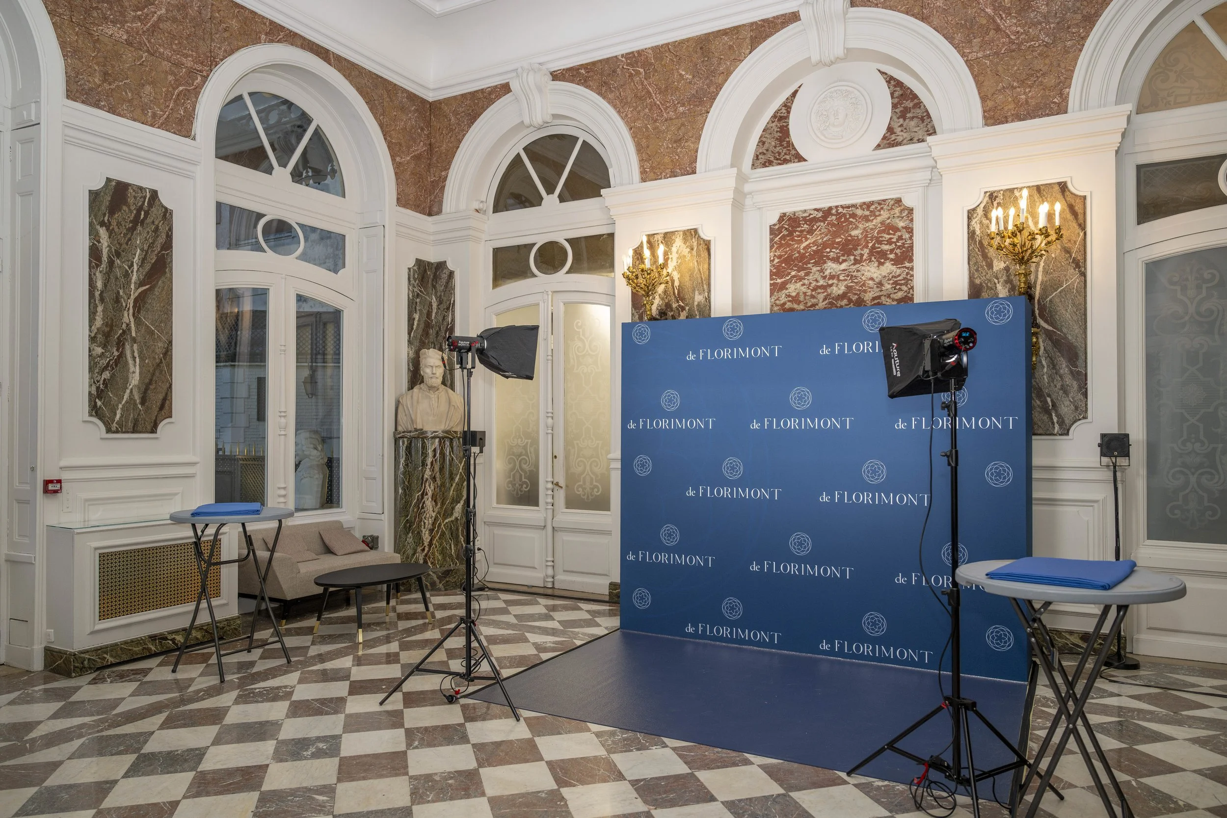 Espace intérieur avec murs ornés de marbre et de moulures blanches, décoré avec une statue et deux lampes en or, comprenant un fond bleu avec le logo de Florimont, deux lampes de studio, deux tables à repassage avec plans de travail.