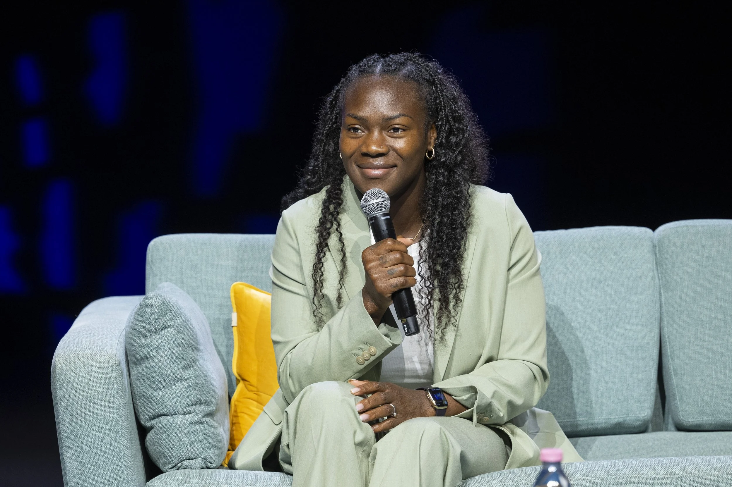 Femme noire habillée en costume clair, assise sur un canapé, tenant un microphone lors d'une discussion ou d'une interview.