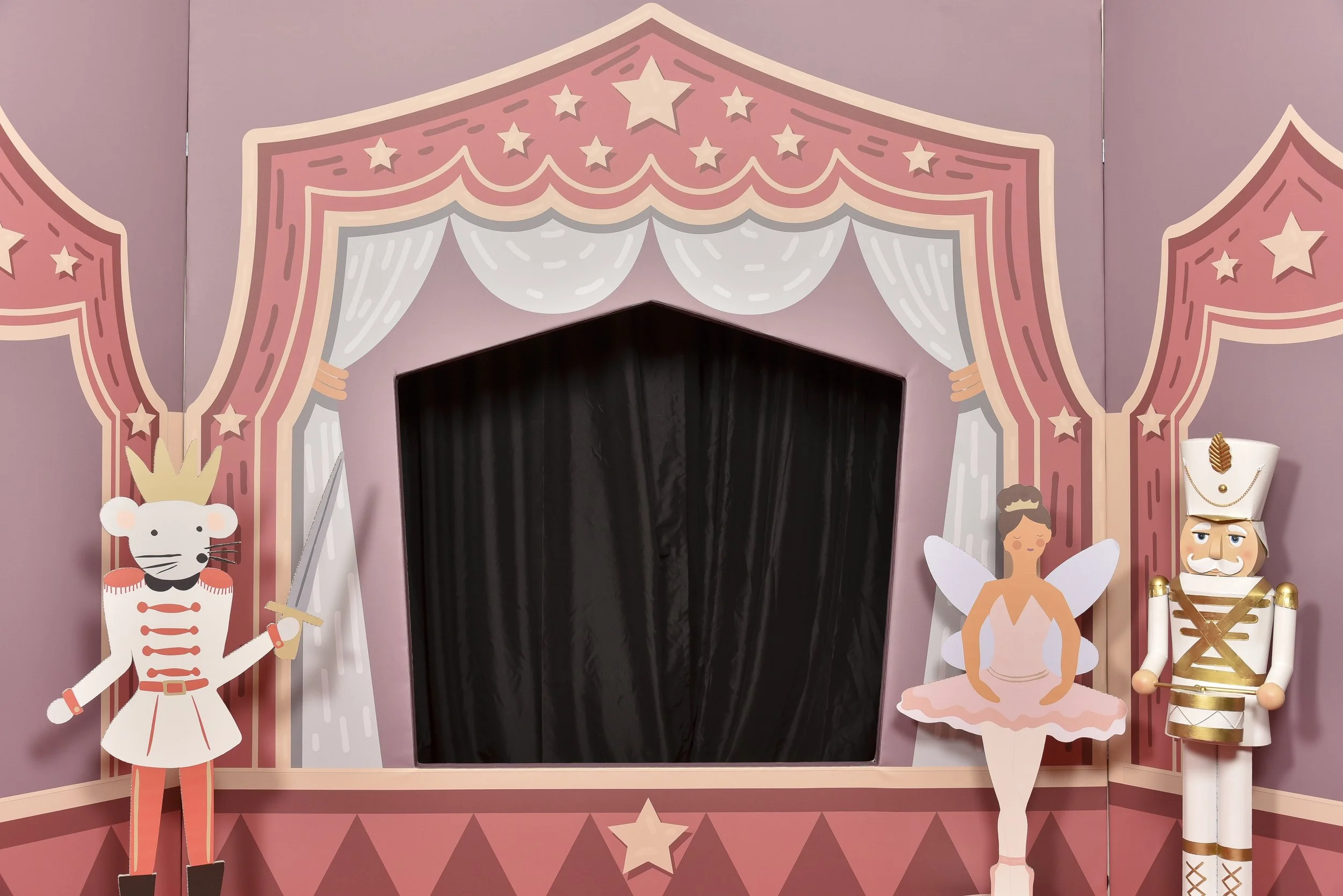 Décor de théâtre en bois avec un rideau noir au centre, entouré de murs peints en rose avec des motifs de étoiles et de draperies blanches. À gauche, un soldat en bois avec une épée, un galet stylisé avec une couronne et moustache. À droite, une fée 