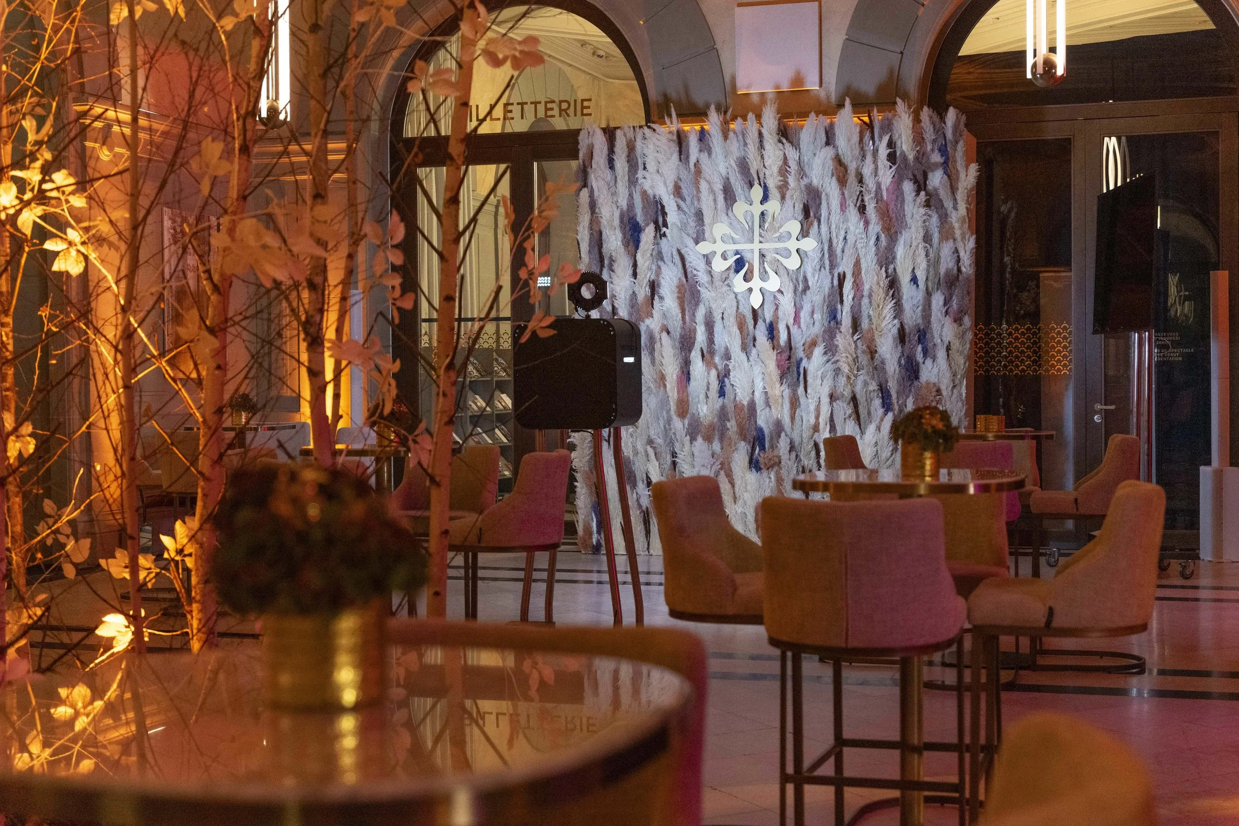 Un intérieur élégant avec des tables rondes, des fauteuils en tissu beige, et une décoration chaleureuse avec un mur en arrière-plan recouvert de plumes, éclairage doux et une décoration de Noël avec un flocon en métal blanc suspendu.