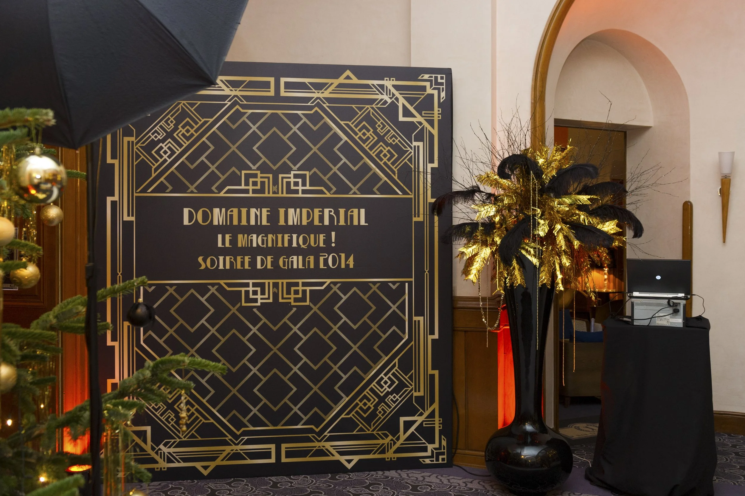 Affiche d'événement élégante pour la soirée de gala 2014 du Domaine Imperial, décorée avec un arbre de Noël et un arrangement floral doré avec des plumes noires, dans une salle bien éclairée.