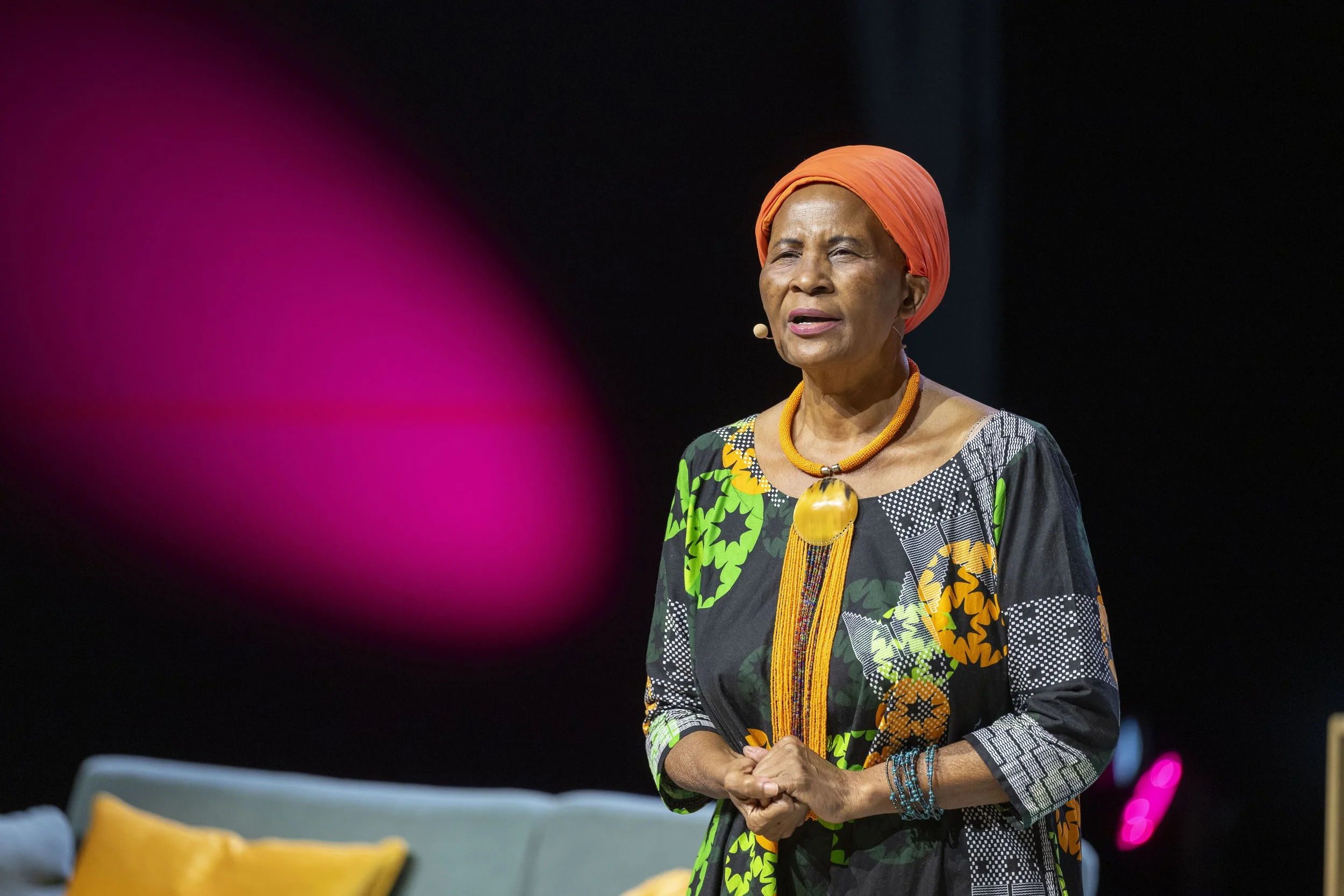 Femme âgée parlant lors d'une présentation, portant un turban orange et un collier coloré, vêtue d'une robe à motifs colorés sur fond noir, sur scène avec un fond sombre.