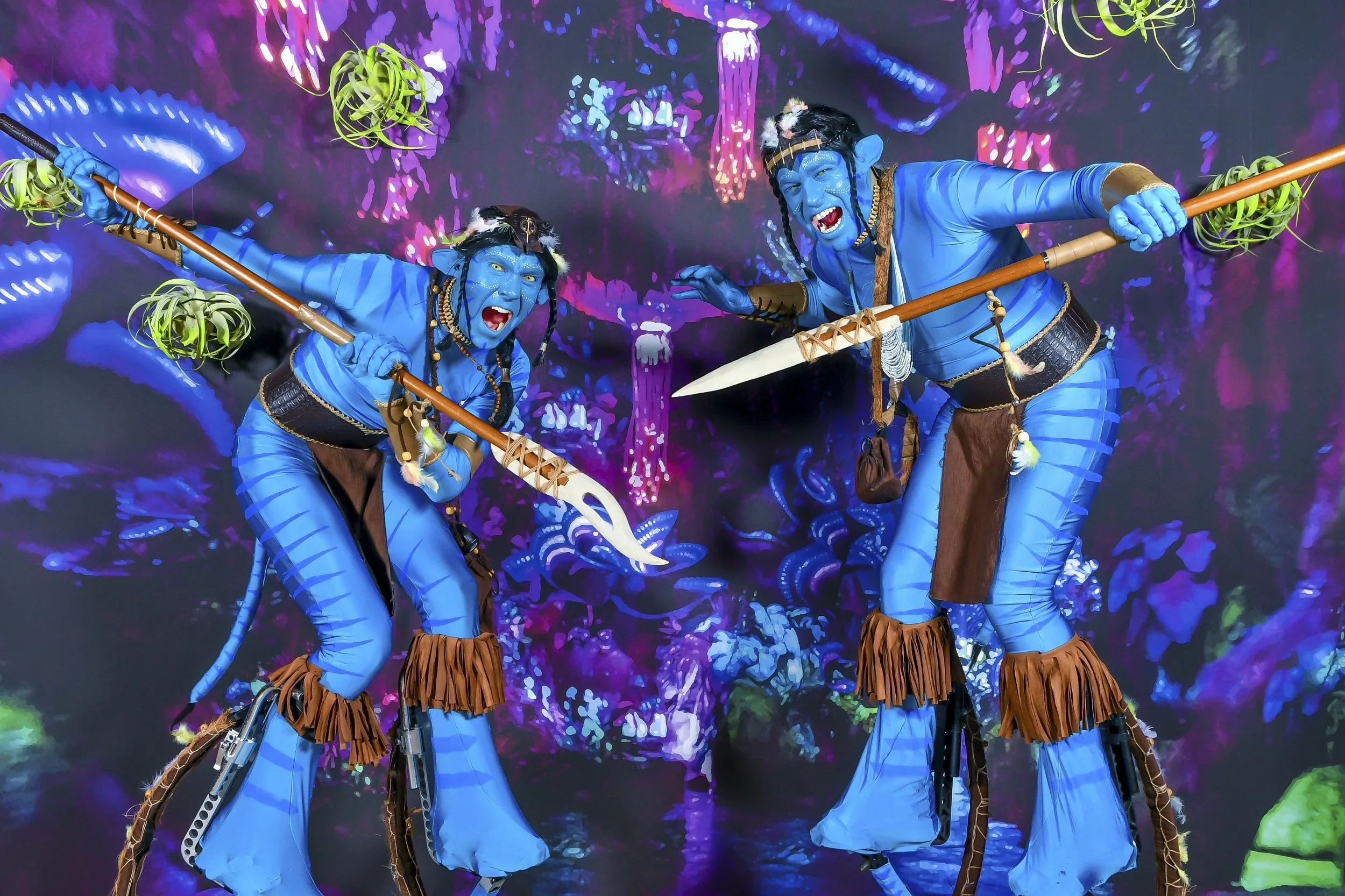 Deux personnes déguisées en personnages de film dans un décor féerique aux couleurs vives, portant des costumes bleus avec des accessoires en cuir et des armes, dans une scène de théâtre ou de fête costumée.