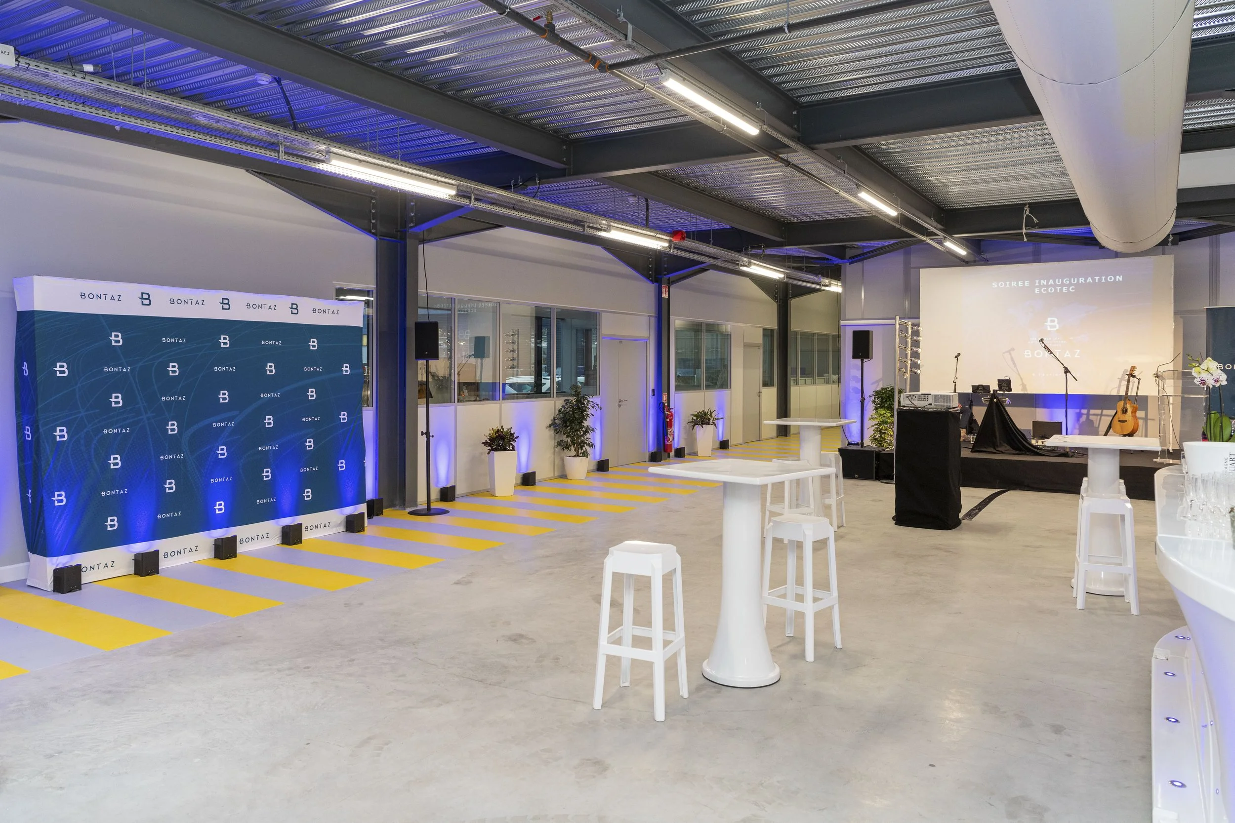 Salle d'événement avec scène, guitare, micros, écran de projection affichant 'SOIREE INAUGURATION ECOTEC', mobilier blanc, plantes, et espace pour invités
