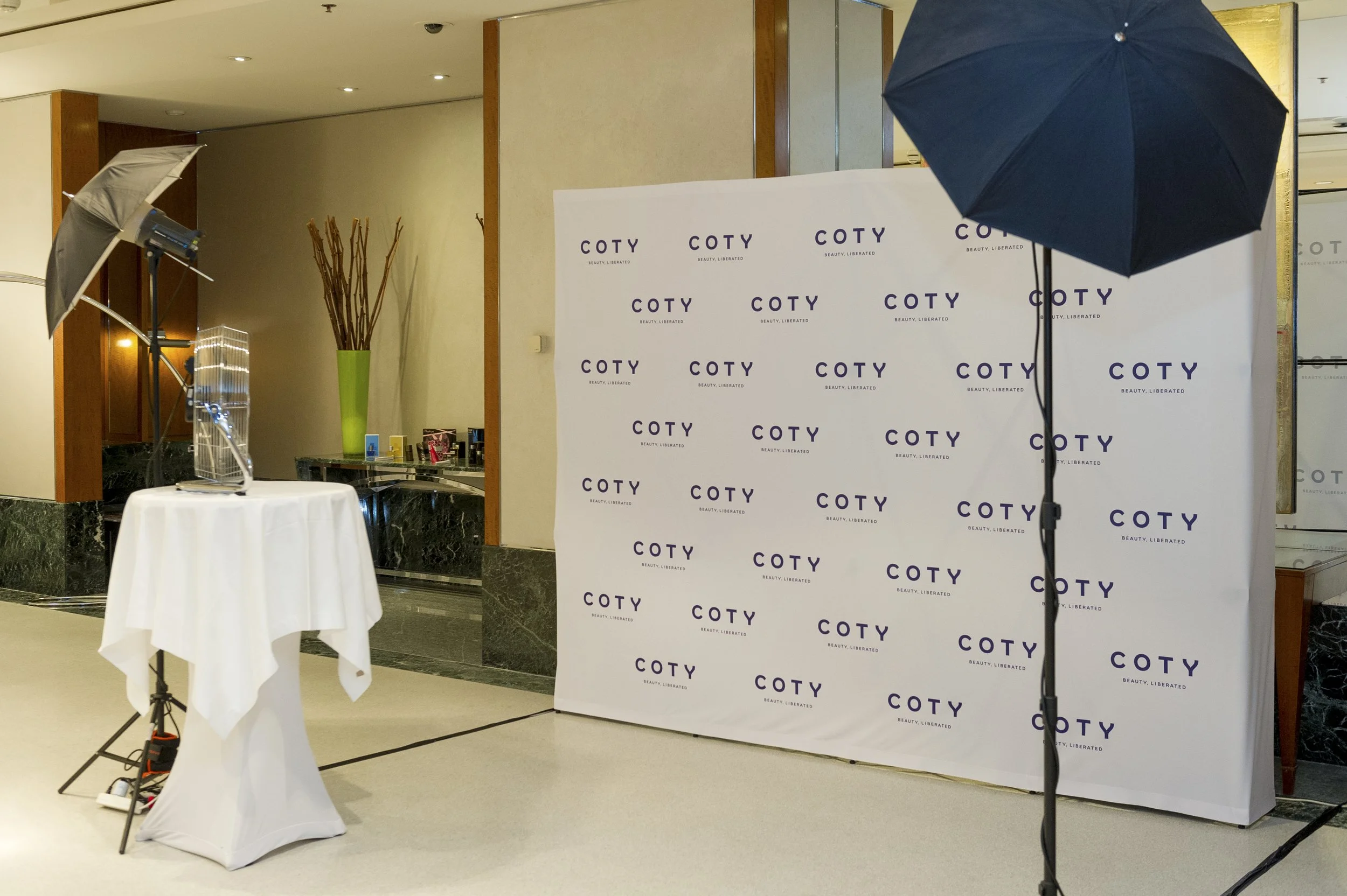 Espace de photographie avec un fond blanc portant le logo 'COTY' et le slogan 'BEAUTY, LIBERATED'. Deux parapluies éclairants, un contre un support avec un drap blanc et quelques équipements photo, ainsi qu'un vase vert avec des branches, décorent l'