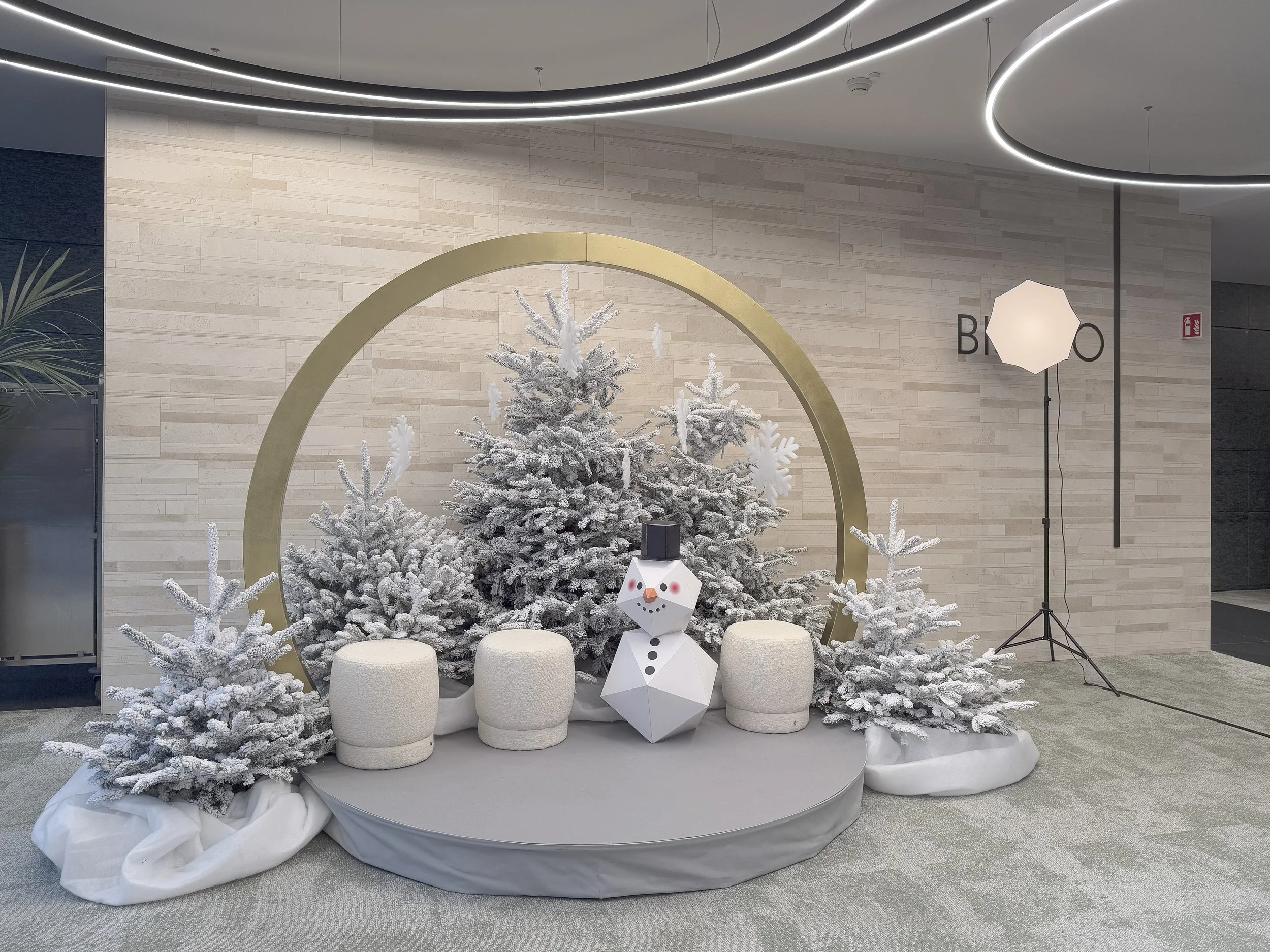 Décor de Noël avec des arbres enneigés artificiels, un bonhomme de neige en papier, un cercle doré, des sièges blancs, et un éclairage de studio dans un espace intérieur.