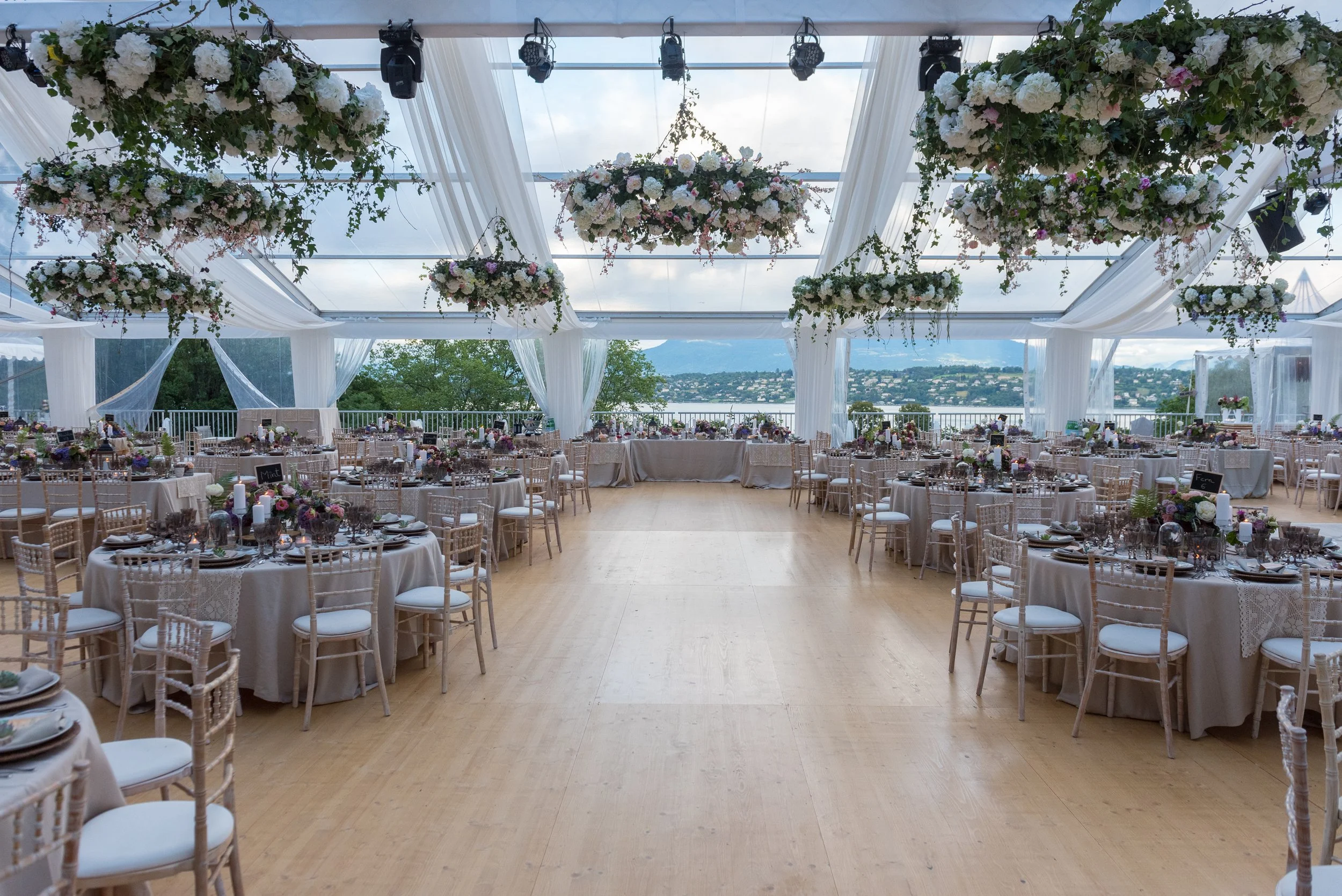 Salle de mariage décorée avec des tables rondes, des chaises en bois, des centres de table floraux suspendus avec des rideaux blancs et une vue sur un lac et des montagnes en arrière-plan.