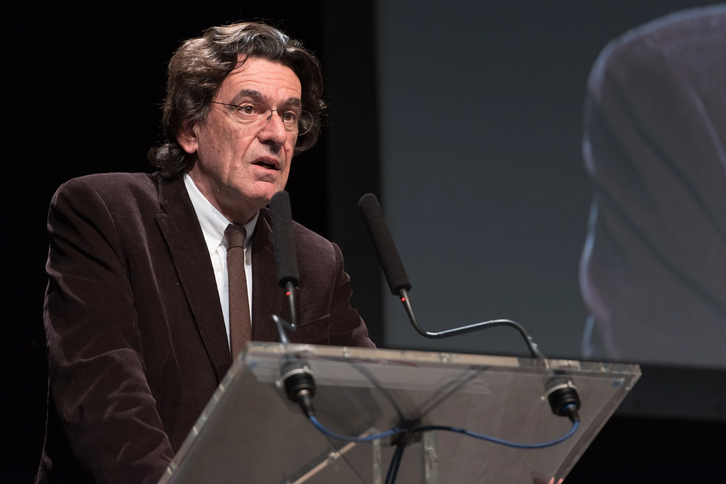 Un homme âgé avec des cheveux gris et ondulés, portant une veste marron, parle à un micro lors d'une conférence ou d'un discours, avec un grand écran derrière lui.