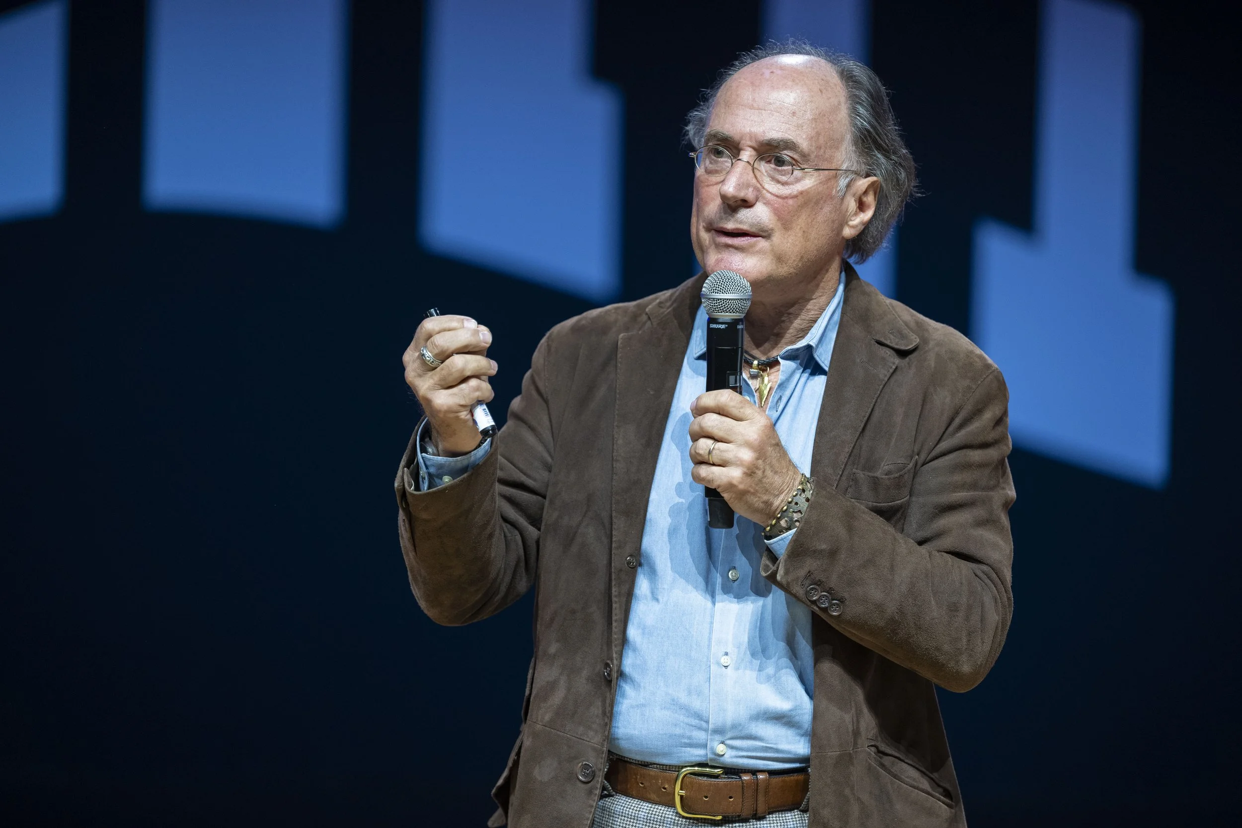 Un homme parlant lors d'une conférence, tenant un micro dans une main et un pointeur dans l'autre, portant une veste en cuir marron et une chemise bleue, contre un fond sombre avec des formes géométriques bleues.