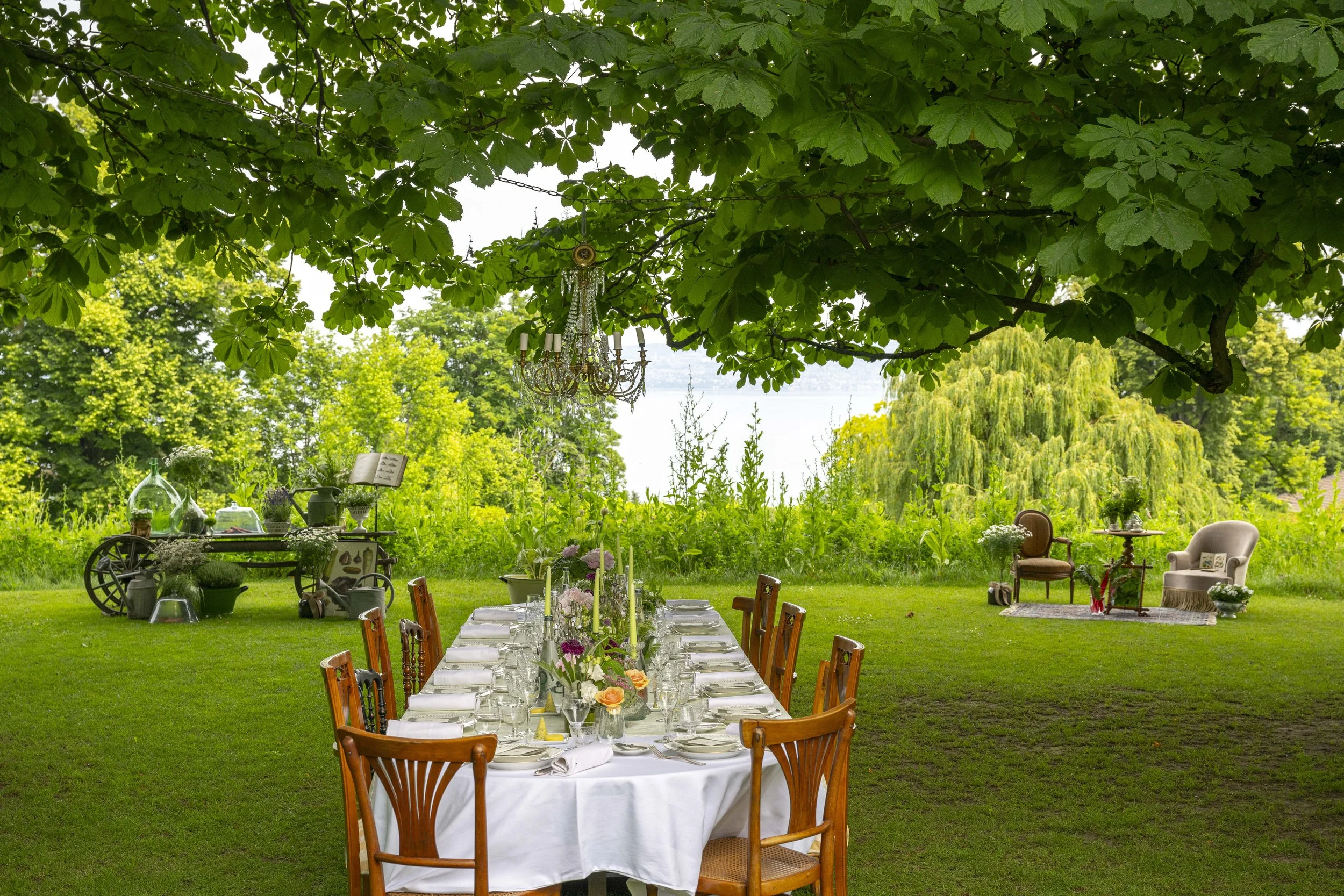 Une table dressée pour un repas en plein air dans un jardin avec des chaises en bois, des fleurs, des bougies et un chandelier suspendu, entourée d'arbres verts.