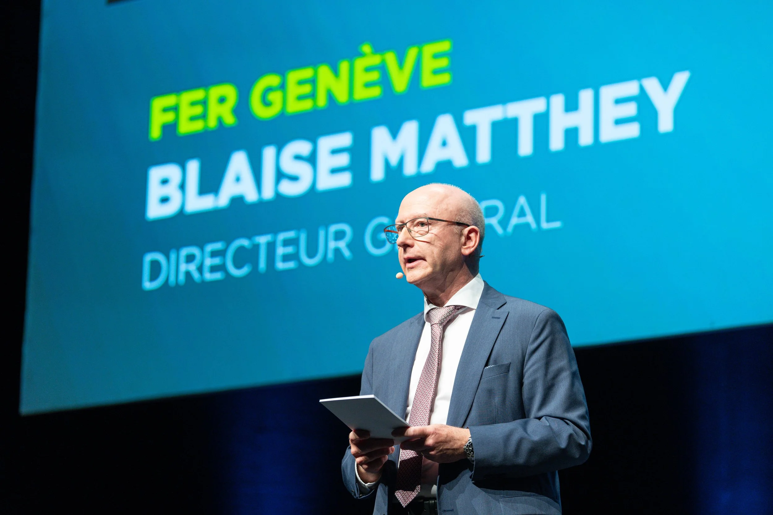 Un homme en costume sur scène avec un écran derrière lui affichant les noms "Fer Genève" et "Blaise Matthey", titulaires de rôles.»