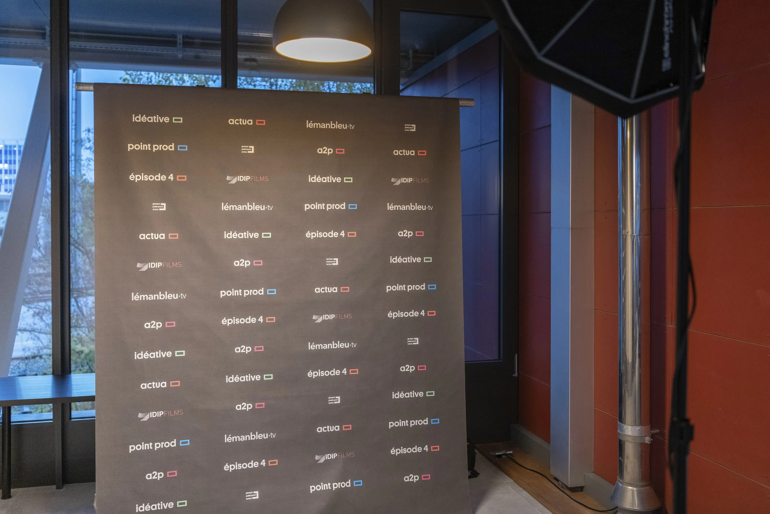 Panneau noir avec plusieurs logos et noms de médias ou programmes, affiché dans une salle intérieure avec fenêtres en arrière-plan et éclairage suspendu.