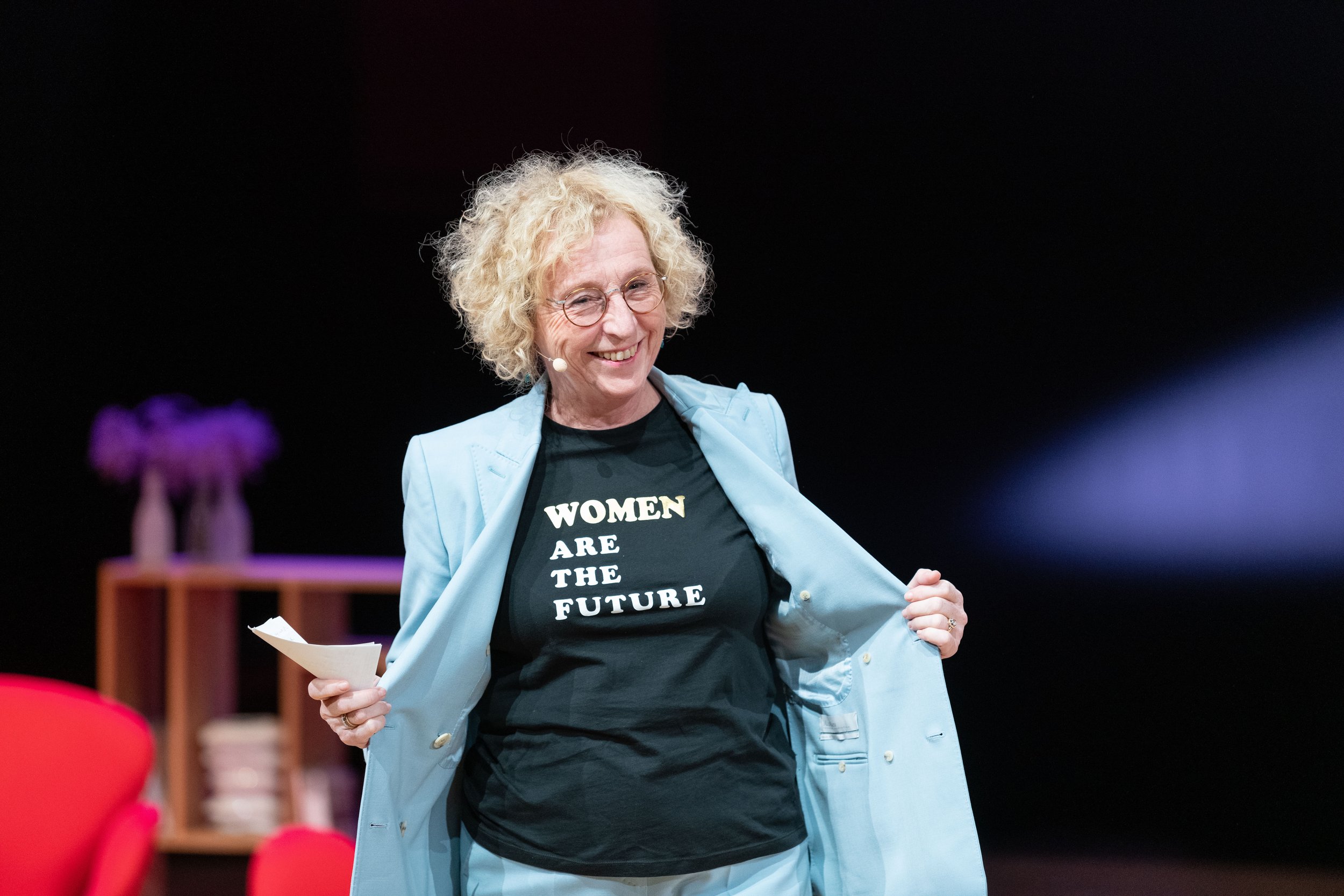 Une femme souriante portant un t-shirt noir avec le message "WOMEN ARE THE FUTURE" et une veste bleue, lors d'une intervention sur scène