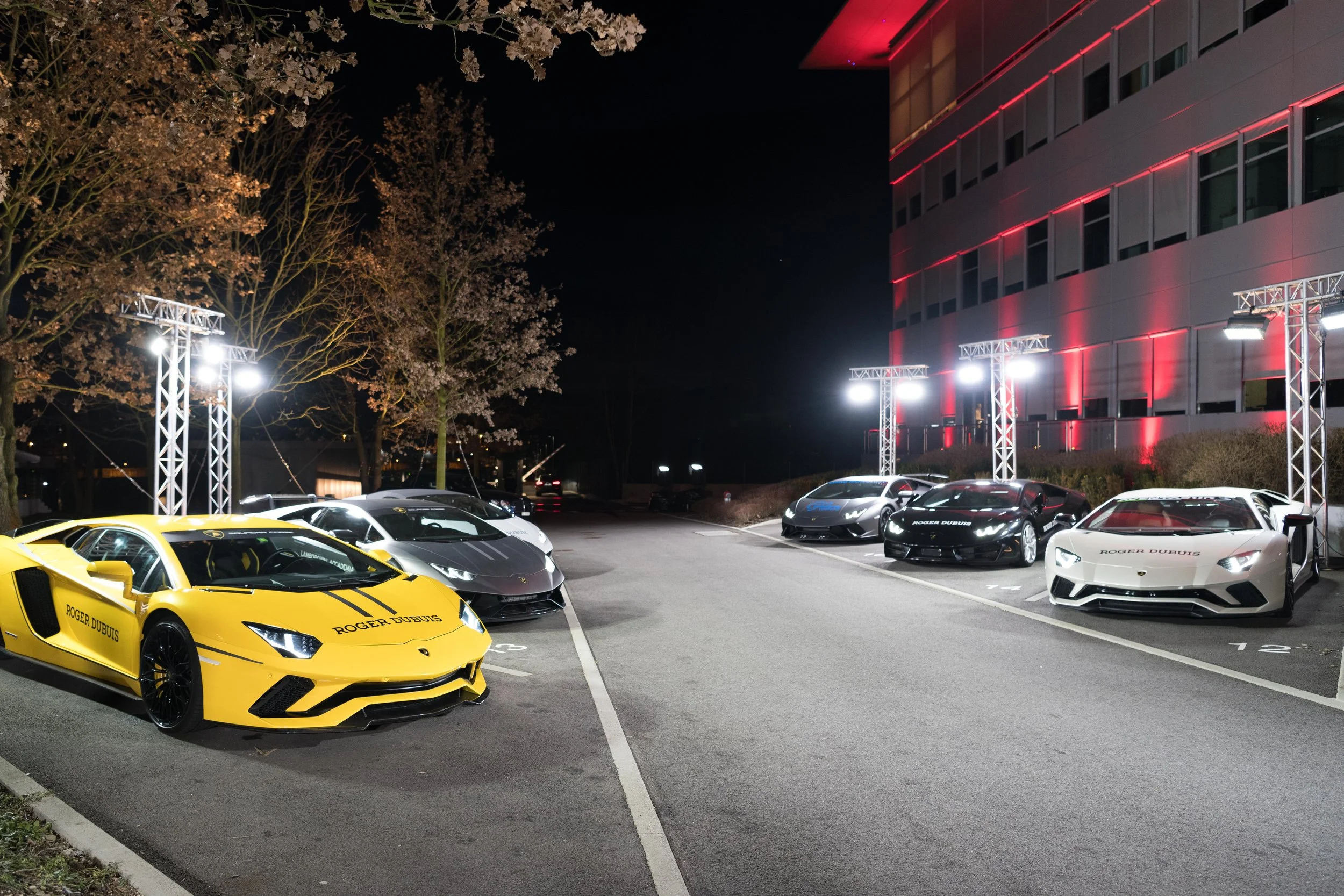 Une rangée de voitures de luxe, notamment plusieurs Lamborghini, garées la nuit devant un bâtiment moderne avec des éclairages. Un éclairage puissant illumine la scène.