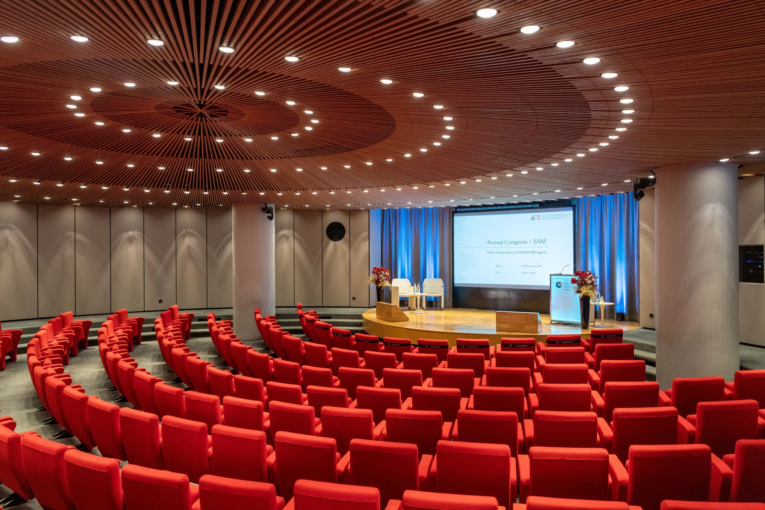 Salle de conférence avec des rangées de sièges rouges, un écran numérique à l'avant avec un podium, des fleurs, et des rideaux bleus, pour un congrès annuel.