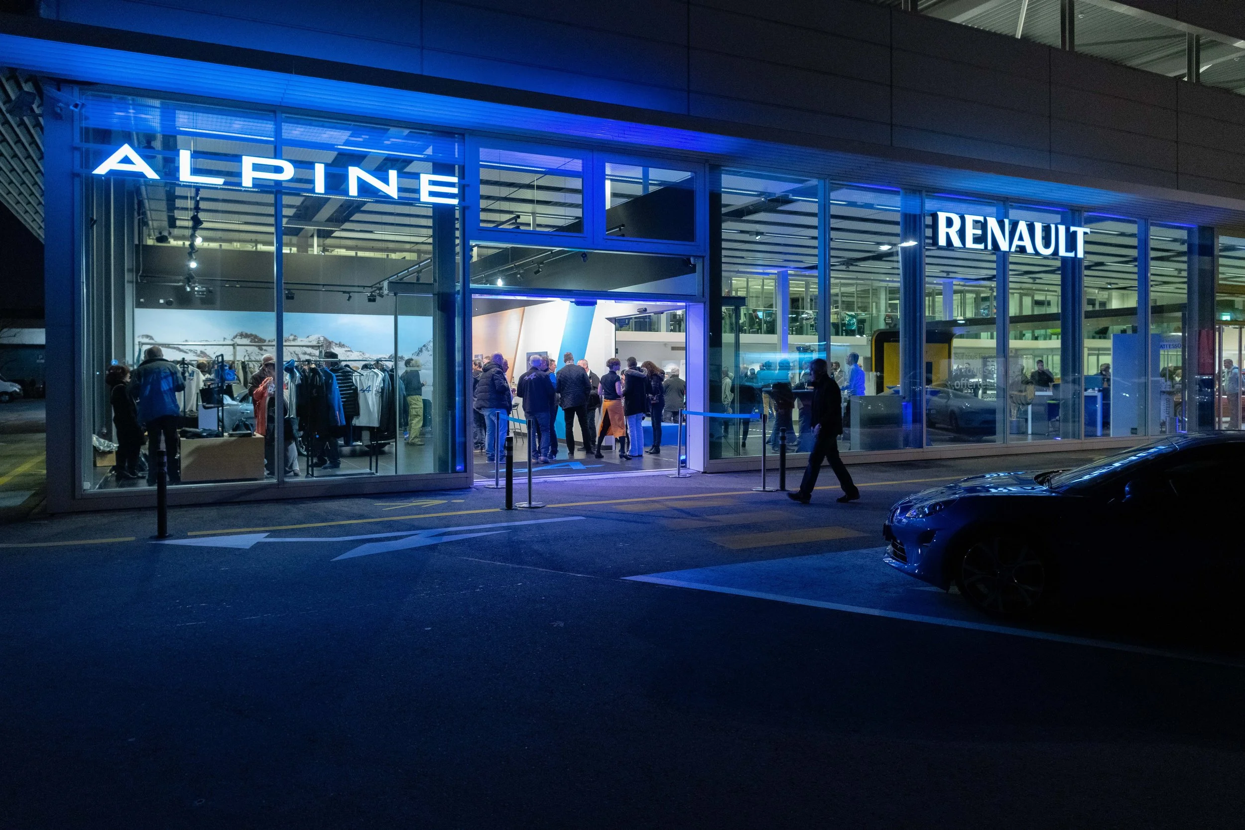 Vitrine d'un magasin avec des néons bleus, portant les noms ALPINE et RENAULT, avec des personnes à l'intérieur et à l'extérieur, soirée ou nuit.