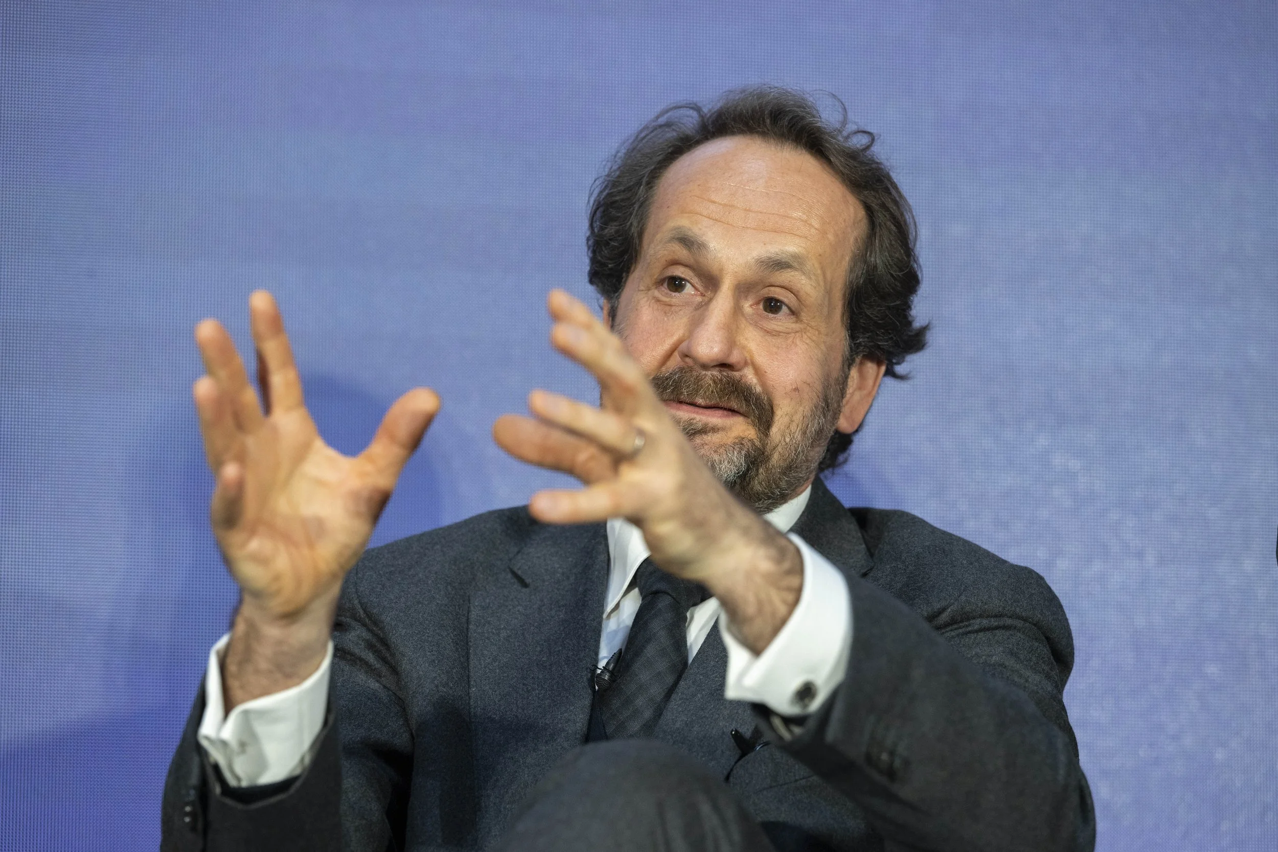 Un homme en costume noir blanc et une cravate, avec des cheveux bruns, parle lors d'une interview ou d'une conférence.