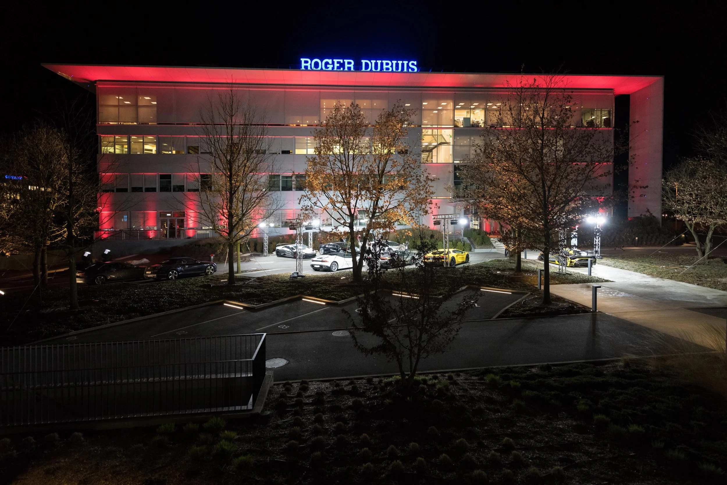 Bâtiment moderne avec enseigne lumineuse 'Roger Dubuis' au sommet, éclairé la nuit, avec parking devant et arbres dénudés.
