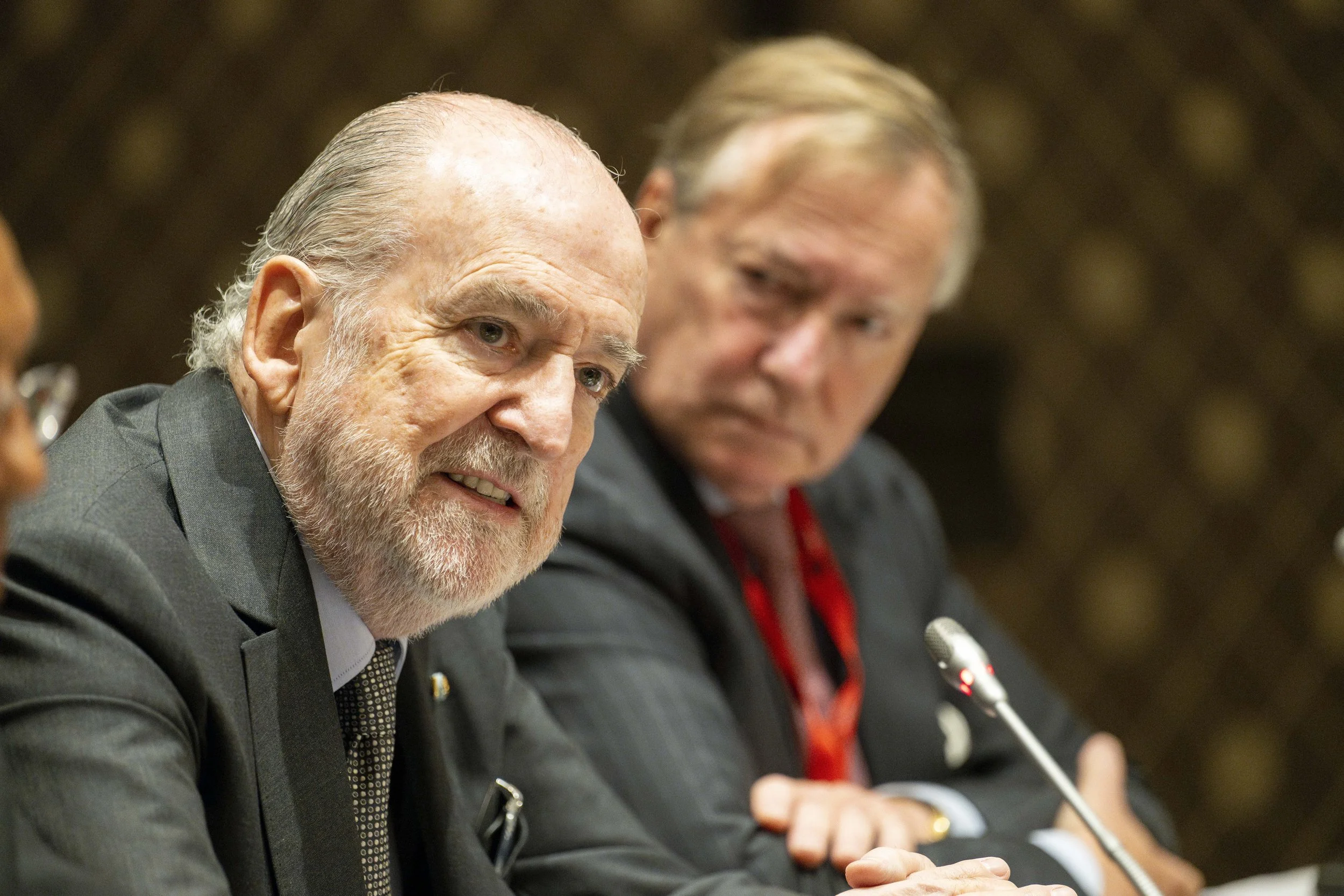 Deux hommes en costume assis à une table lors d'une réunion ou conférence, avec un micro devant eux, l'un parlant ou parlant, l'autre regardant.