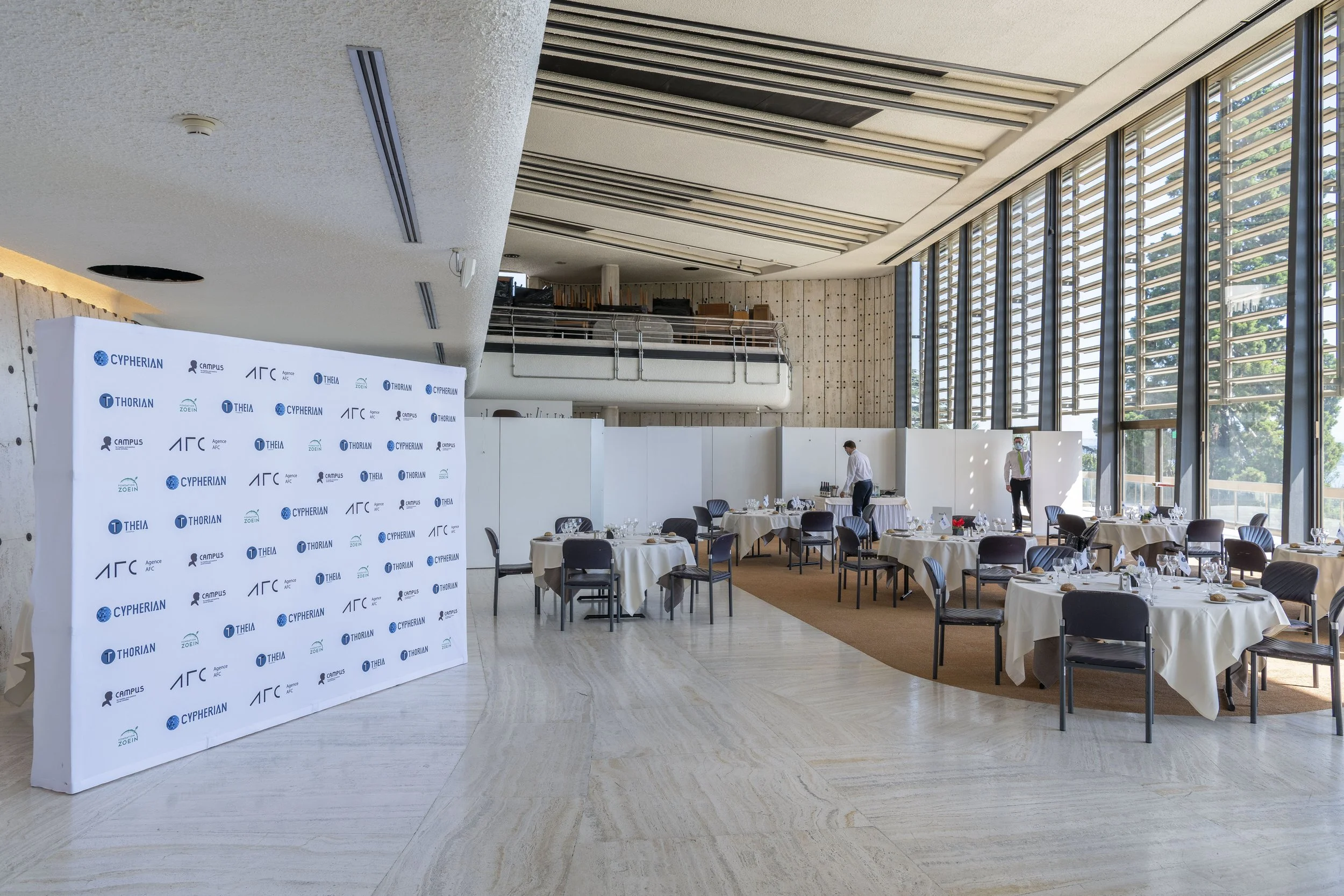 Salle de réception avec plusieurs tables rondes couvertes de nappes blanches, serviettes et vaisselle, deux personnes en train de préparer la salle, grandes fenêtres permettant de laisser entrer beaucoup de lumière naturelle, décor moderne et élégant