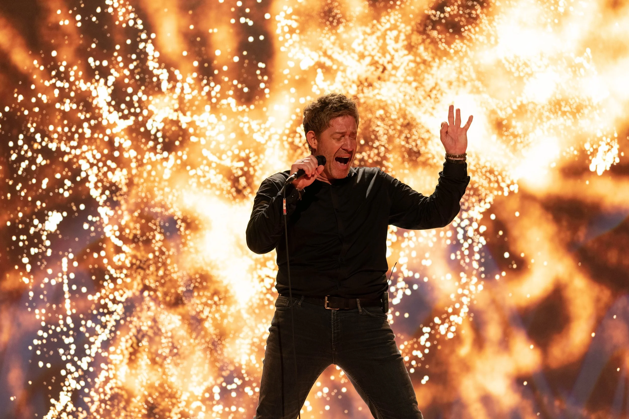 Un homme chante avec passion sur scène, entouré de feux d'artifice en arrière-plan lors d'un spectacle ou concert.