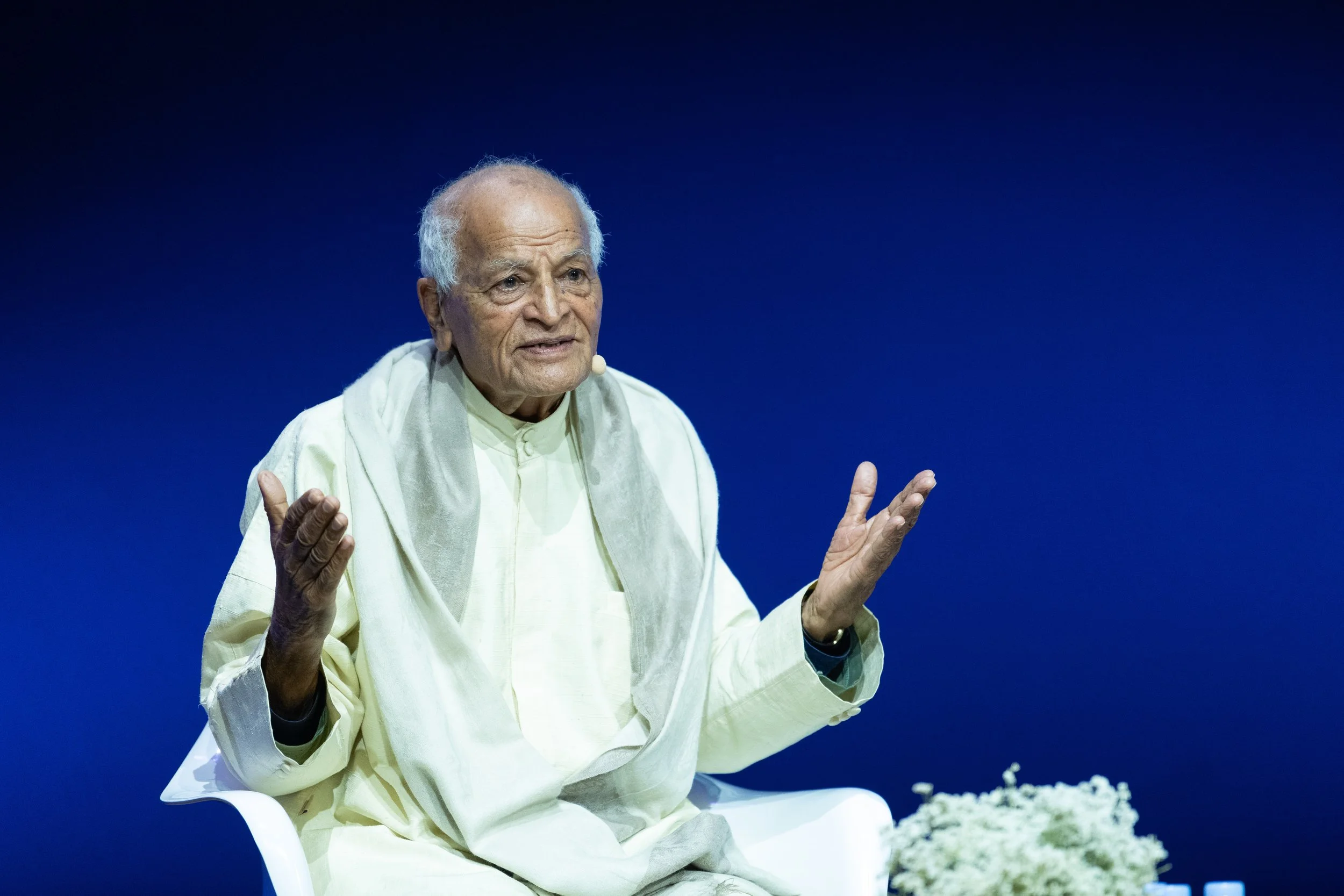 Un homme âgé, vêtu de vêtements traditionnels, assis sur une chaise blanche, parlant ou s'exprimant lors d'une conférence ou d'un discours, avec un fond bleu