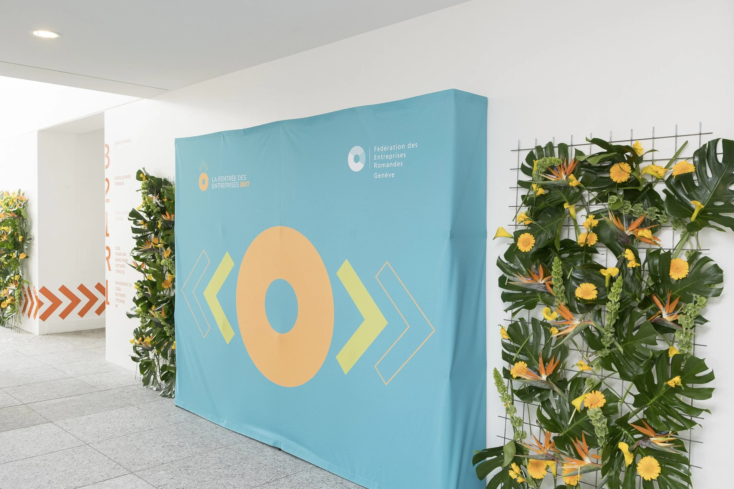 Décor pour une conférence ou un événement avec un panneau bleu clair indiquant 'La rentrée des entreprises 2017' et le logo de la Fédération des Entreprises Romandes Genève, entouré de installations florales vertes et jaunes.