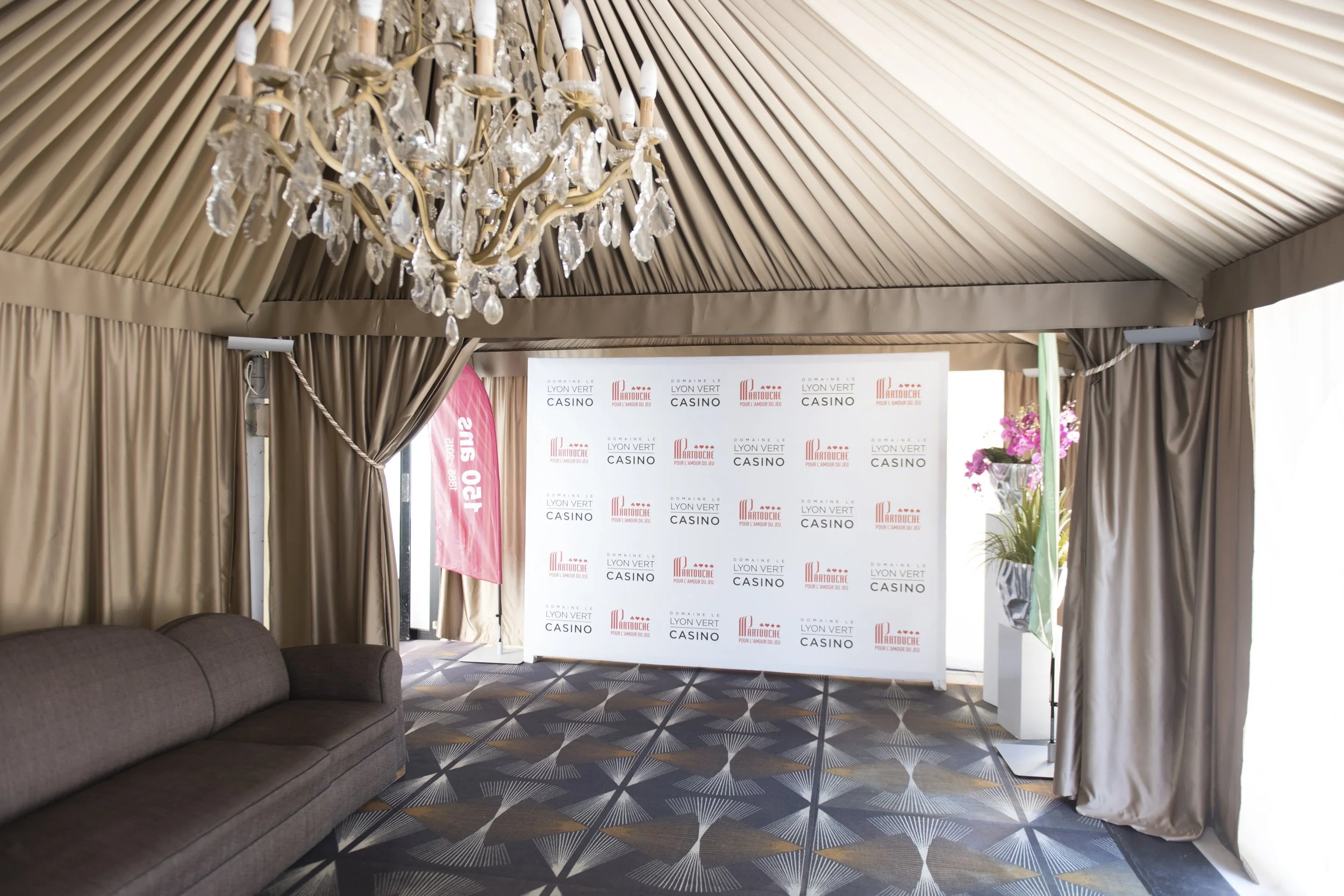 Intérieur d'une tente élégante avec un chandelier en cristal suspendu, un tapis à motifs géométriques, un canapé marron sur la gauche, et un panneau de fond blanc avec logos de casino et de Domaine Le Lyon Vert, ainsi que des fleurs roses dans une po