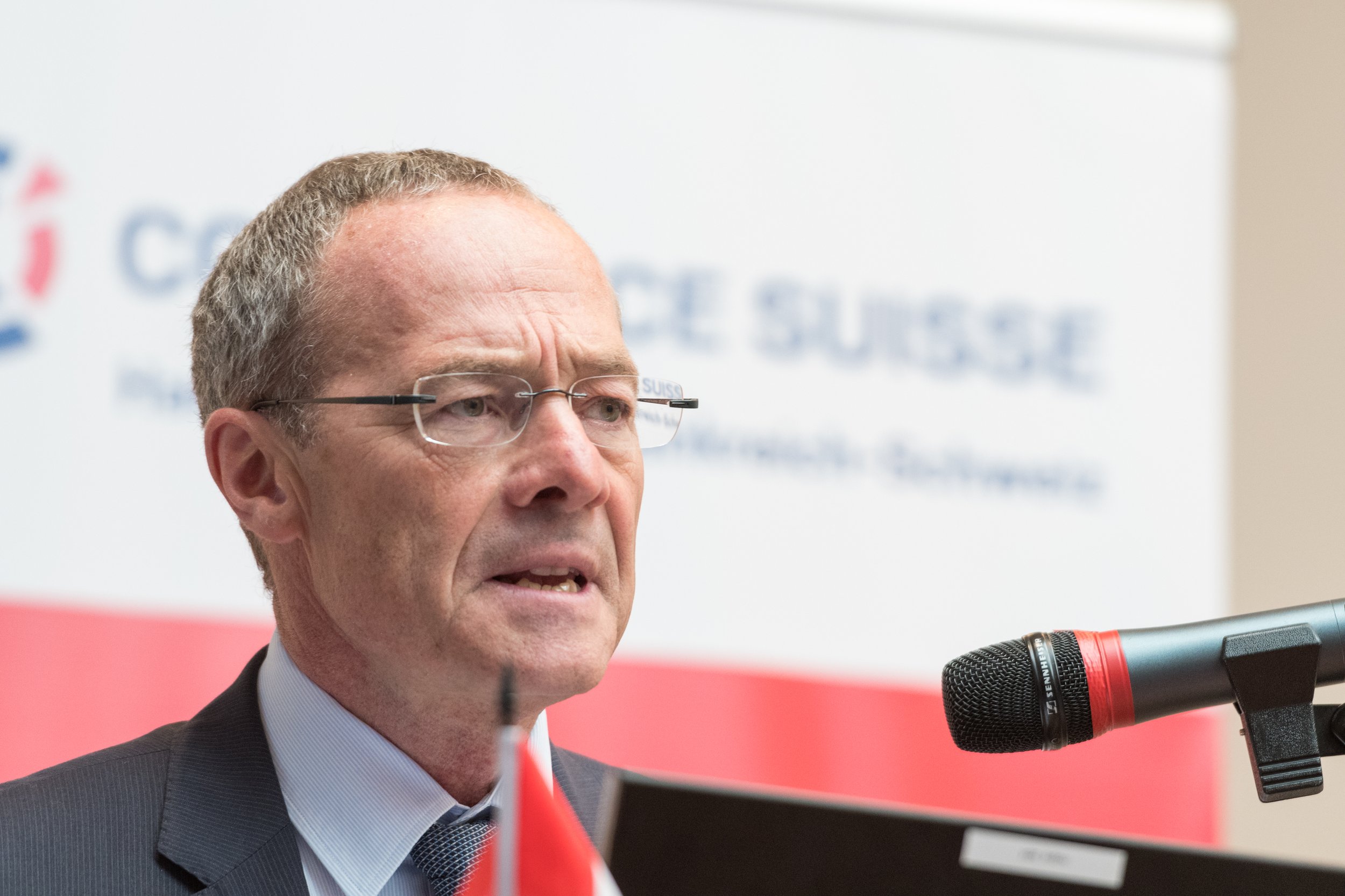 Homme en costume avec lunettes parlant devant un micro lors d'une conférence, avec un drapeau et un logo en arrière-plan.