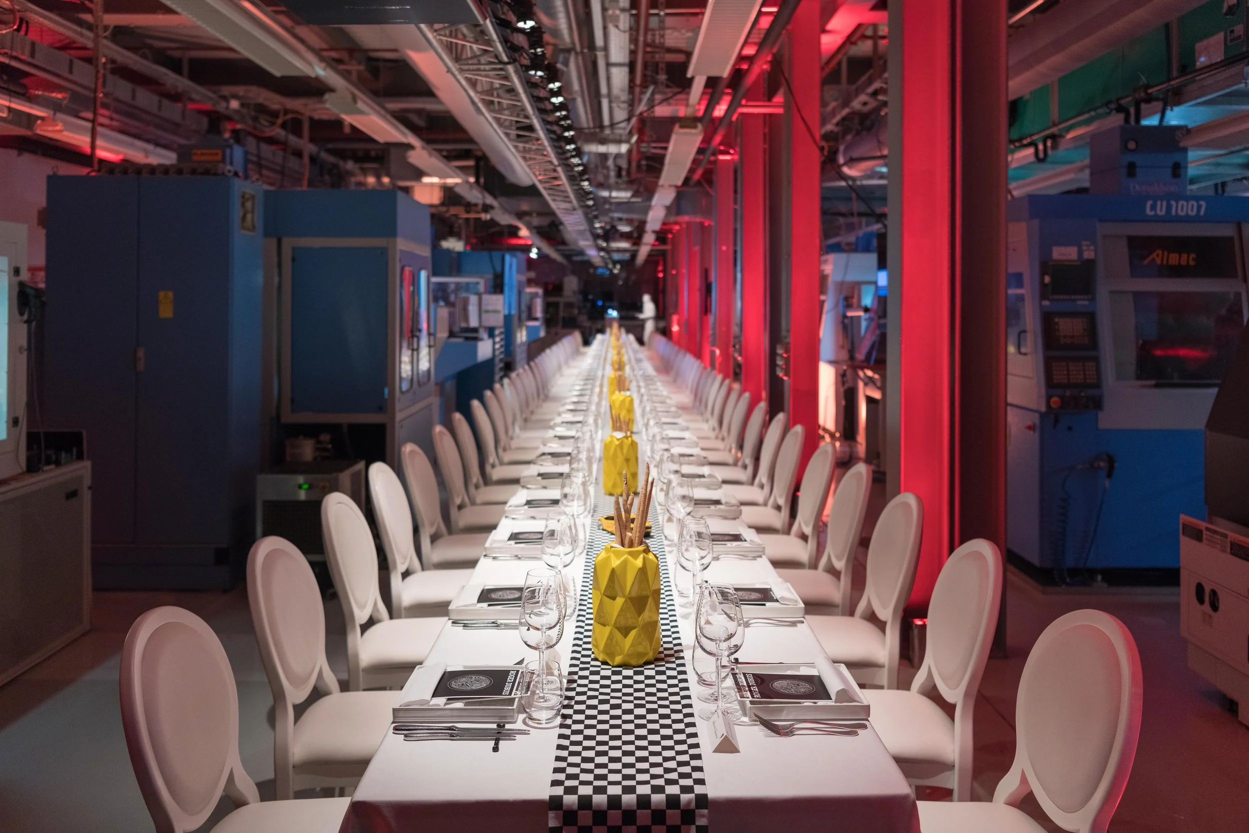 Table de dîner longue dans un environnement industriel avec chaises blanches, décor de vases jaunes, éclairage tamisé rouge et équipements techniques en arrière-plan.