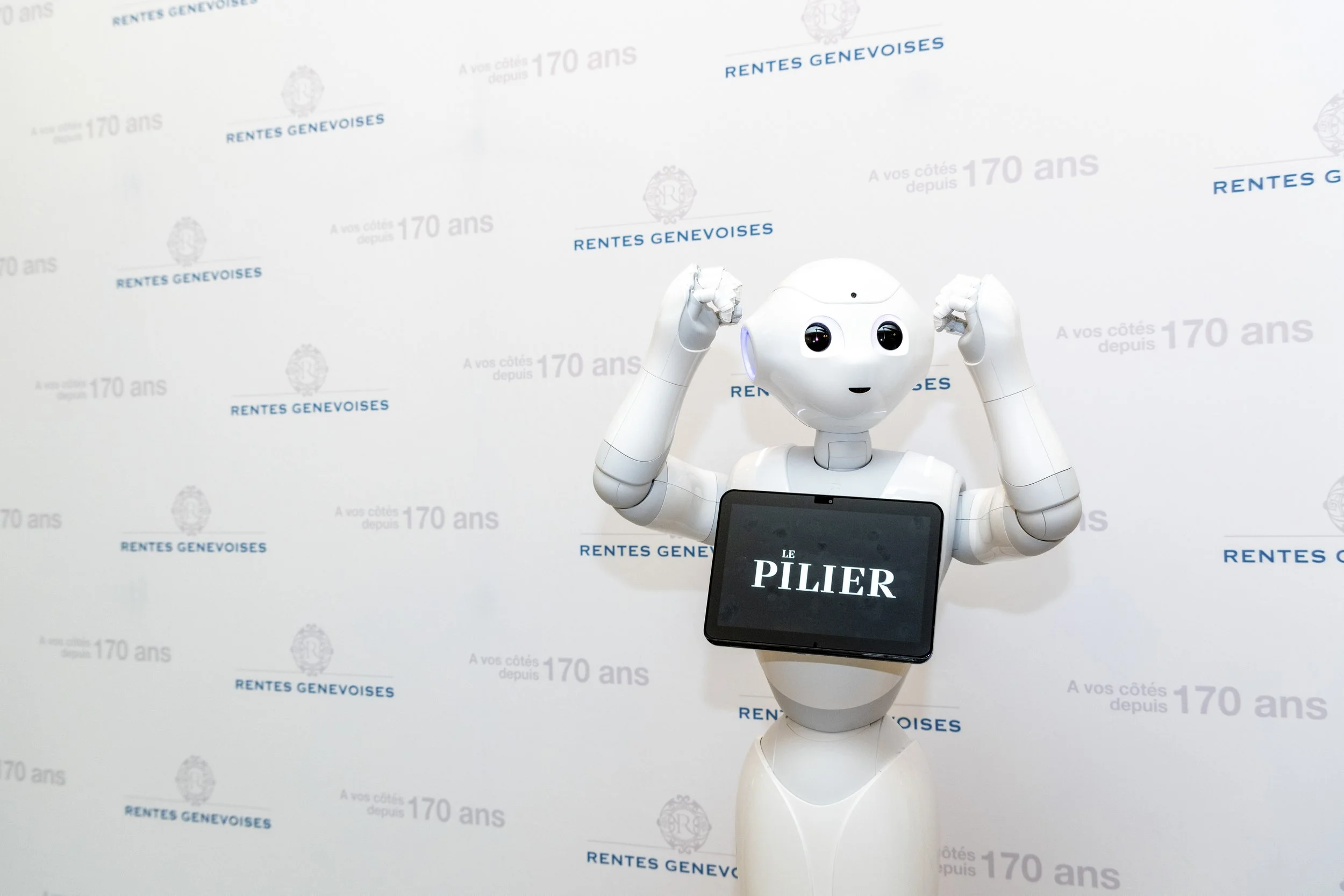 Un robot blanc avec un écran affichant « LE PILIER » sur la poitrine, levant les bras dans une pose de force devant un mur blanc avec du texte en français et en bleu, comprenant des logos et des phrases telles que « RENTES GENEVOISES » et « A vos côt