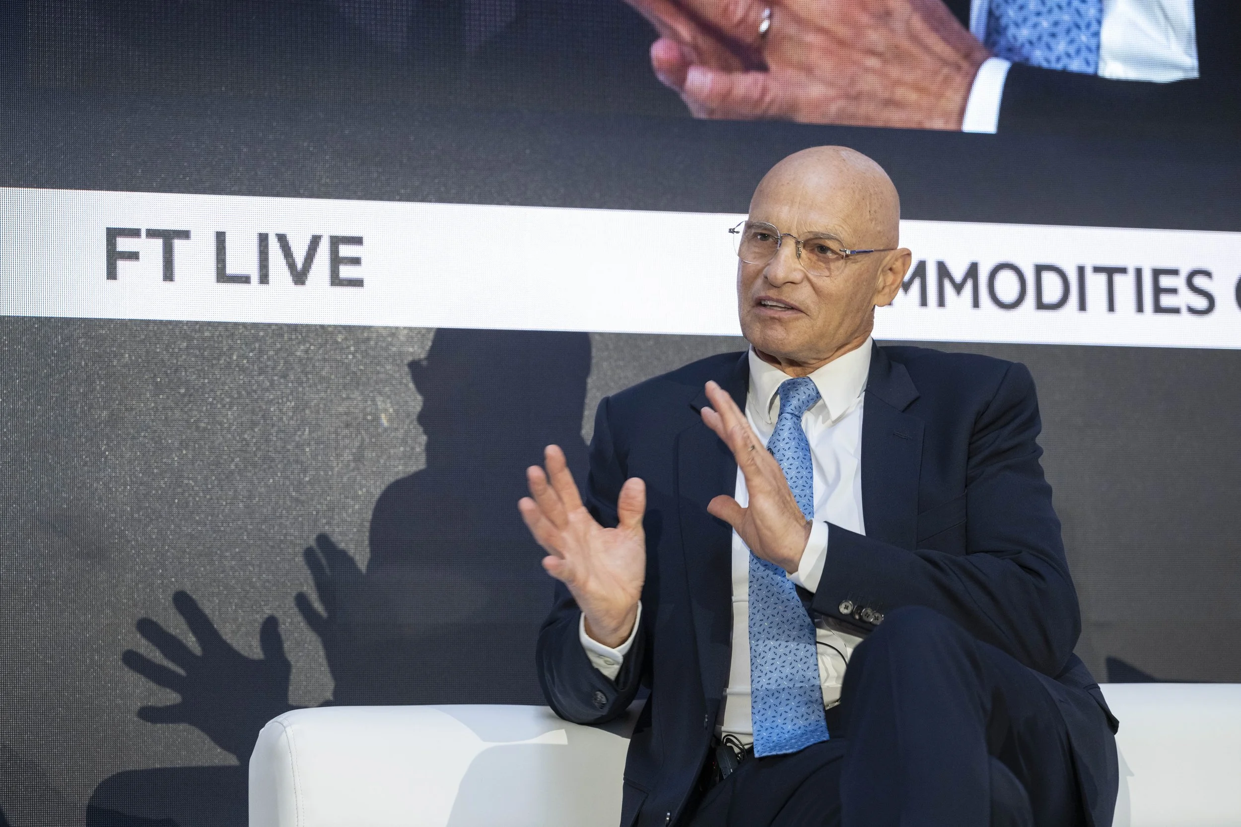 Homme en costume bleu avec une chemise blanche et une cravate bleue parlant lors d'une interview ou conférence, avec un fond affichant les textes "FT LIVE" et d'autres mots en anglais.