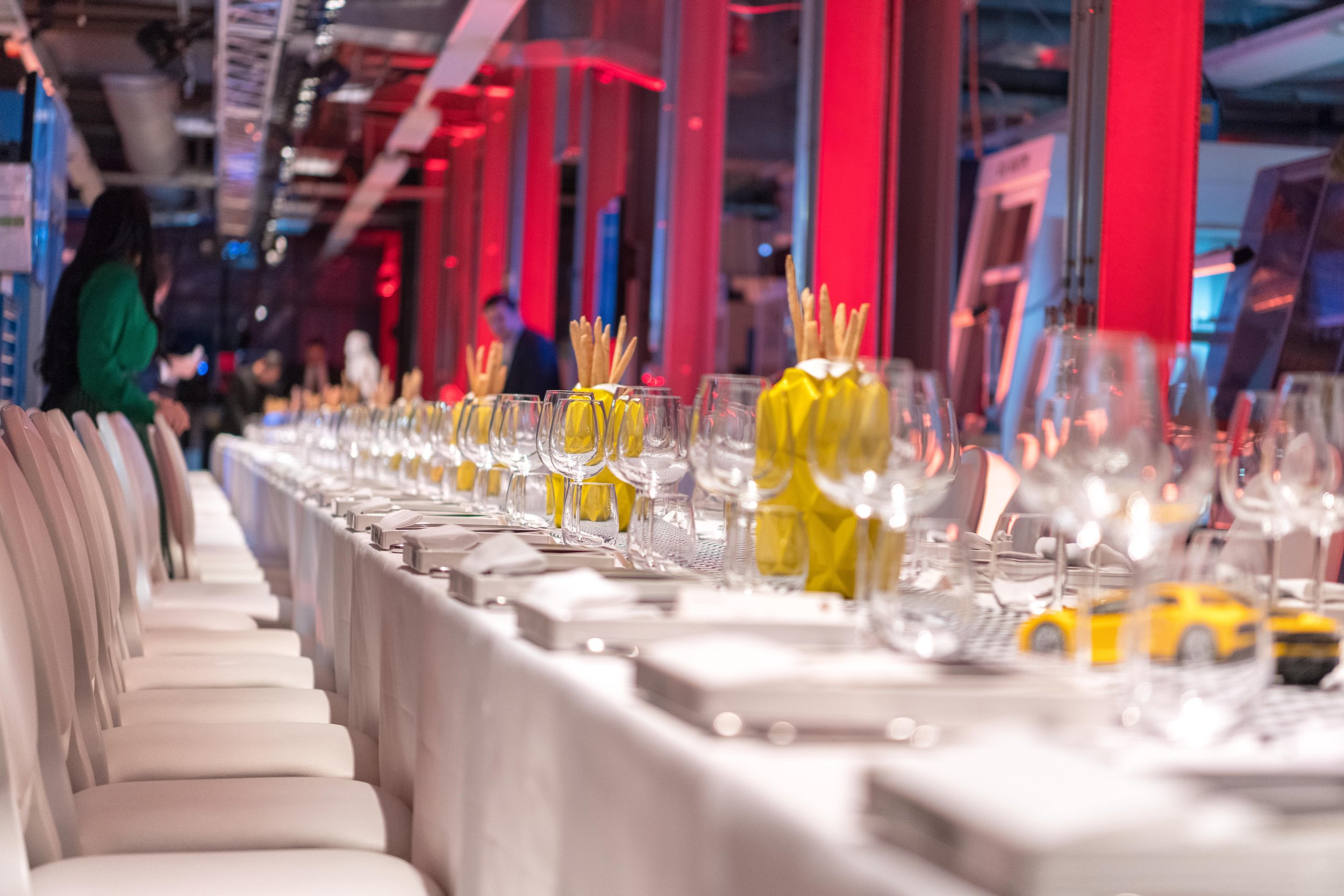 Une longue table élégamment dressée pour un repas, avec des verres, des assiettes, des couverts, des serviettes pliées dans des vases jaunes, et des petits véhicules jaunes comme décoration. L'ambiance est lumineuse avec des éclairages rouges et bleu