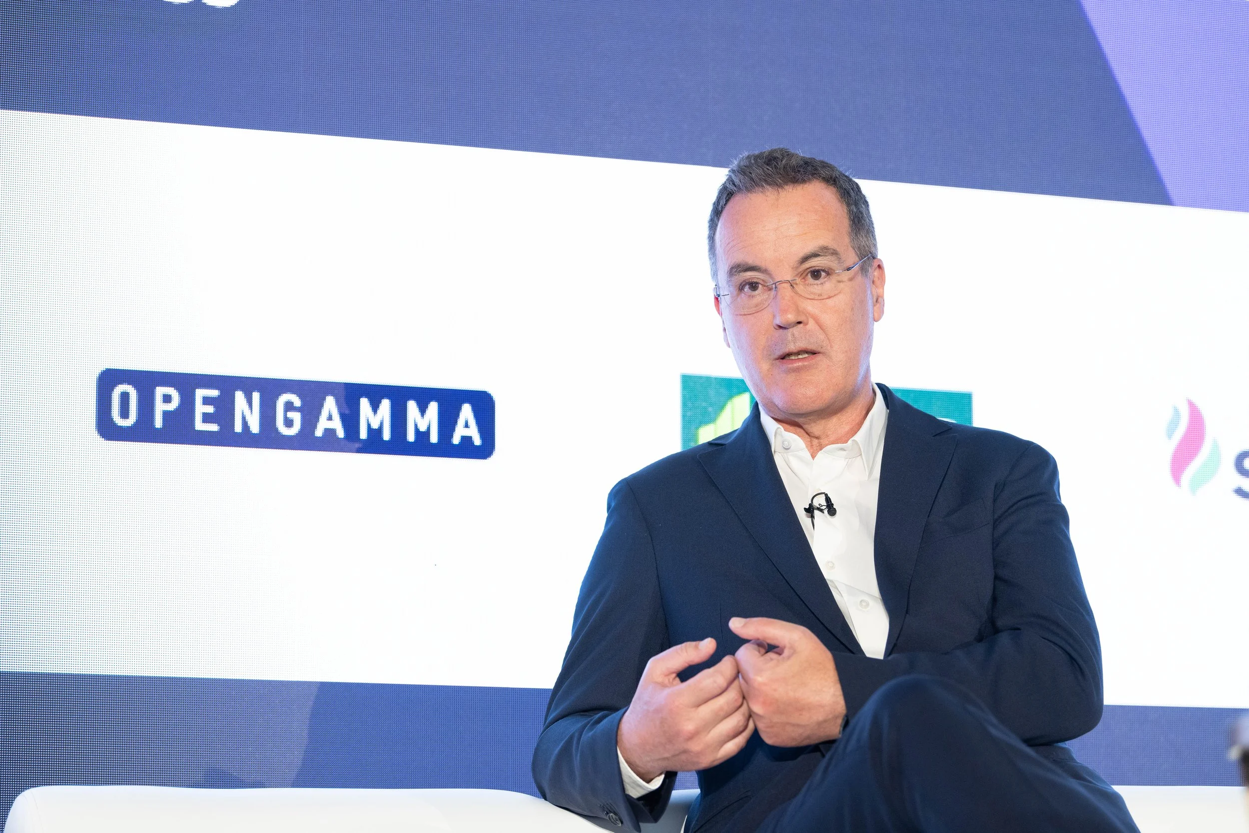 Homme en costume assis lors d'une présentation ou d'une conférence, avec logos de 'OPENGAMMA' visible à l'arrière-plan.