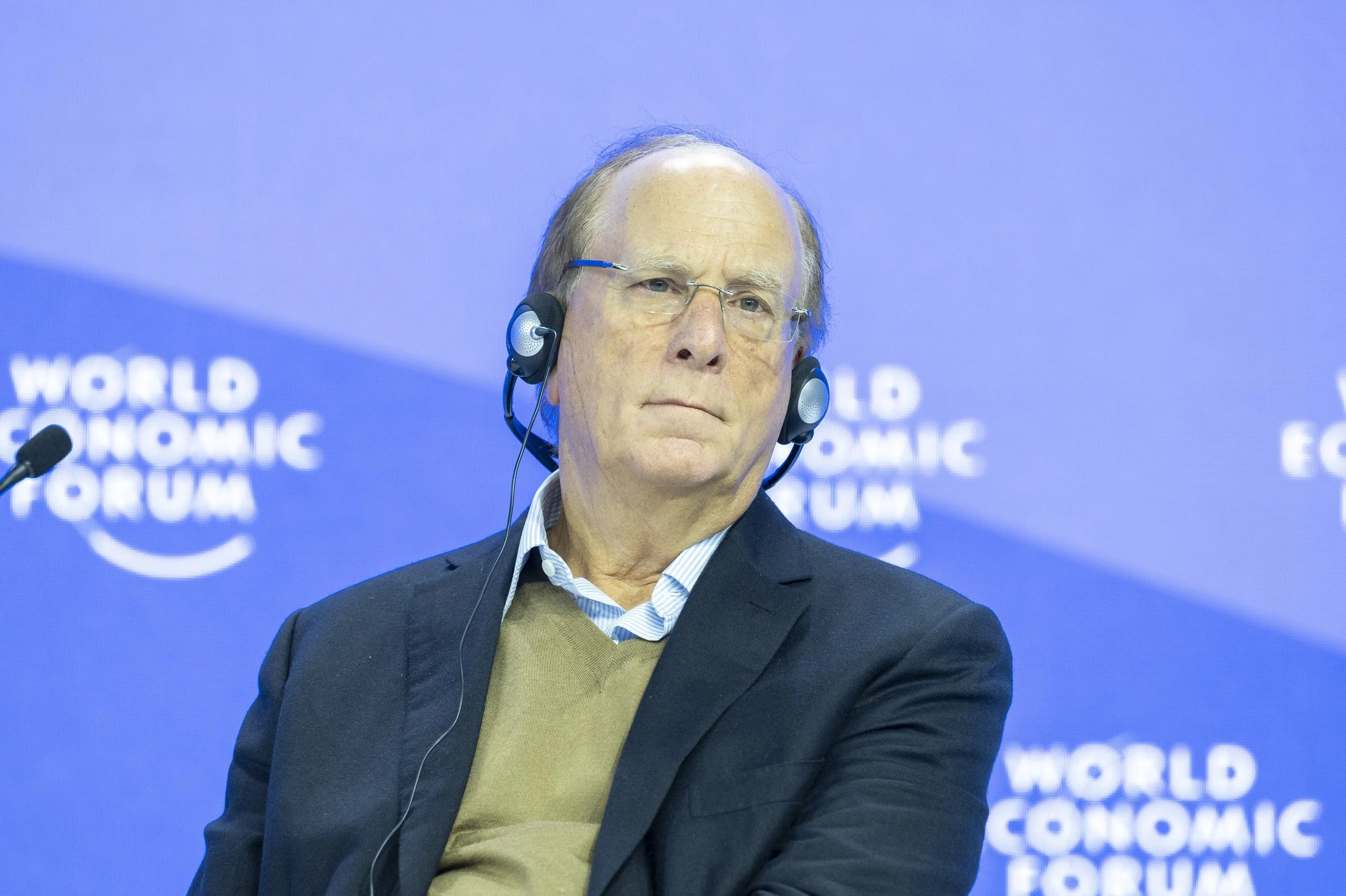 Un homme âgé porte un costume sombre, une chemise à rayures et un pull beige, portant un casque audio en écoutant attentivement lors du Forum Économique mondial.