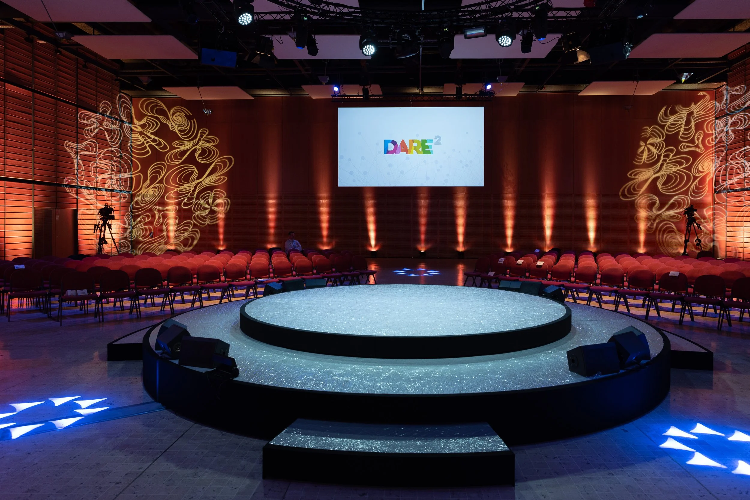 Salle de conférence avec un grand écran à l'avant affichant "DARE²" et une scène ronde éclairée