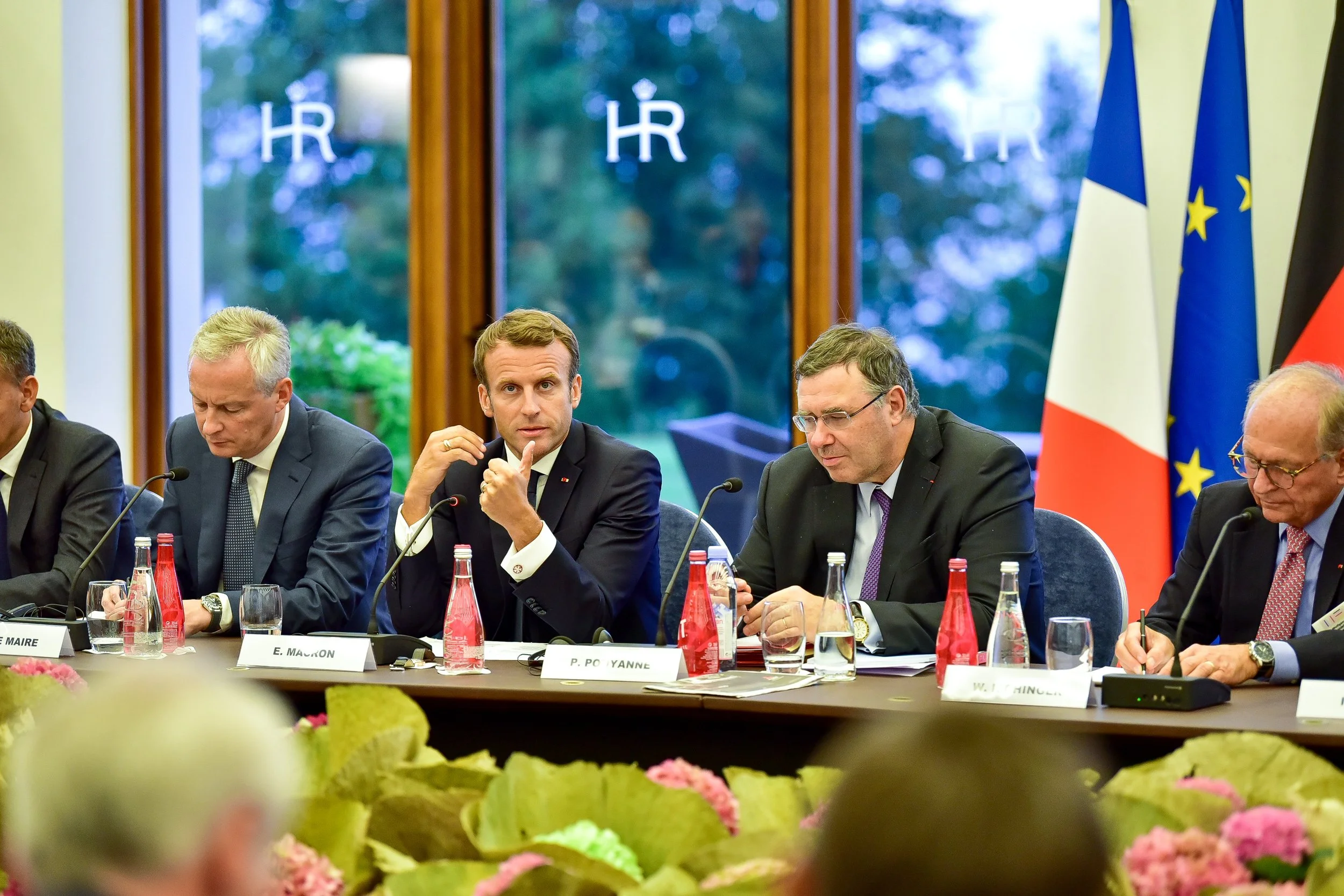 Groupe de sept personnes assises à une table lors d'une réunion officielle, avec drapeaux français, européen et allemand en arrière-plan et micro sur la table.