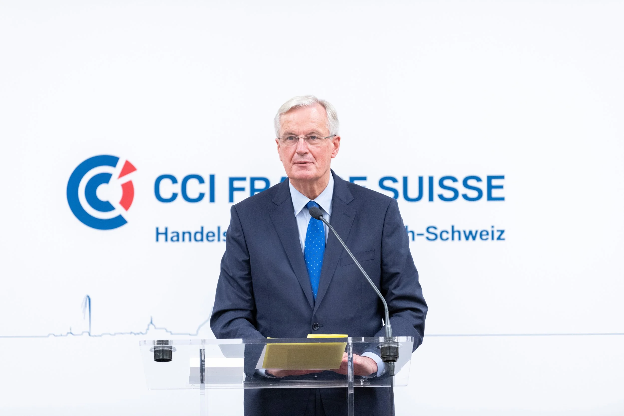 Homme en costume parlant à un micro lors d'une conférence avec le logo de la CCI Suisse sur un fond blanc.