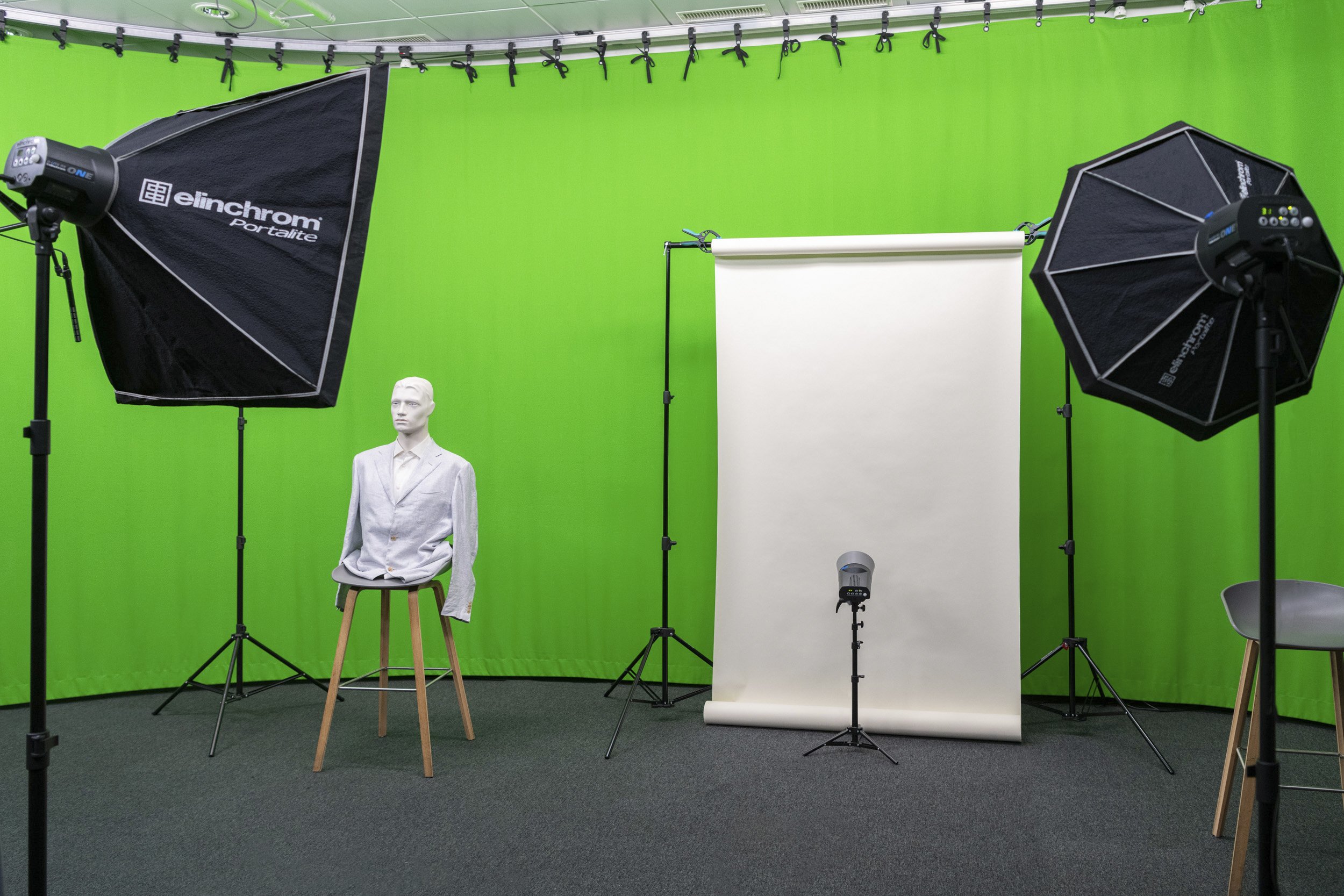 Studio de photographie avec un fond vert, un mannequin vêtu d'un costume gris, des équipements d'éclairage, un panneau de fond blanc, un microphone sur trépied, et une chaise.