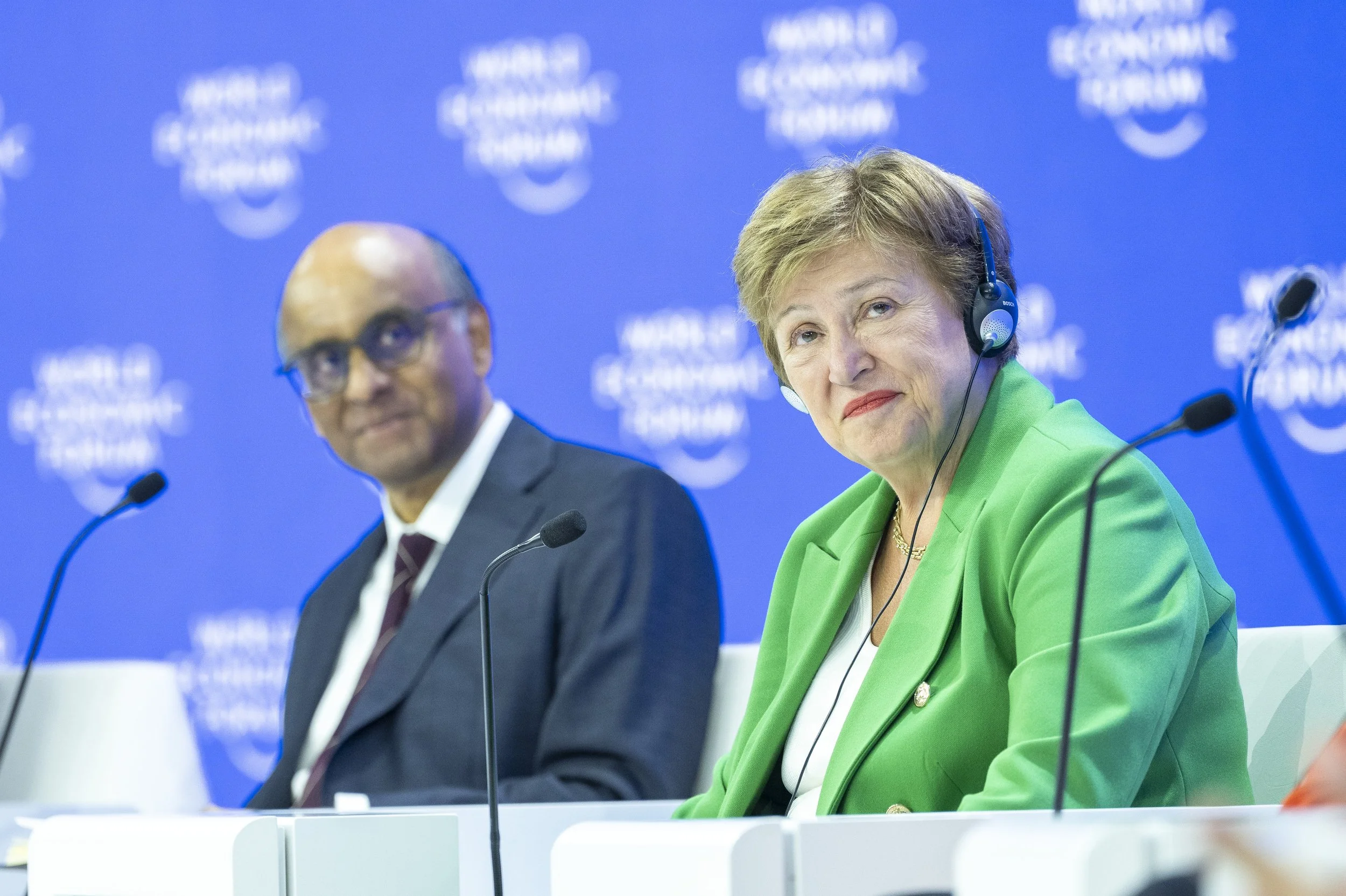 Deux personnes assises lors d'une conférence avec un fond bleu affichant un logo de Forum Économique Mondial. La personne à droite est une femme portant un blazer vert et un casque audio. La personne à gauche est un homme portant un costume sombre, d