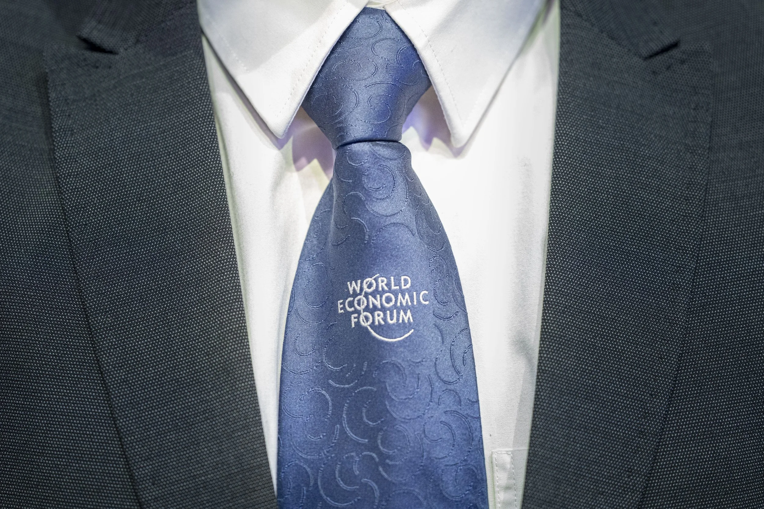 Veste de costume sombre, chemise blanche et cravate bleue avec le logo 'World Economic Forum' imprimé dessus.